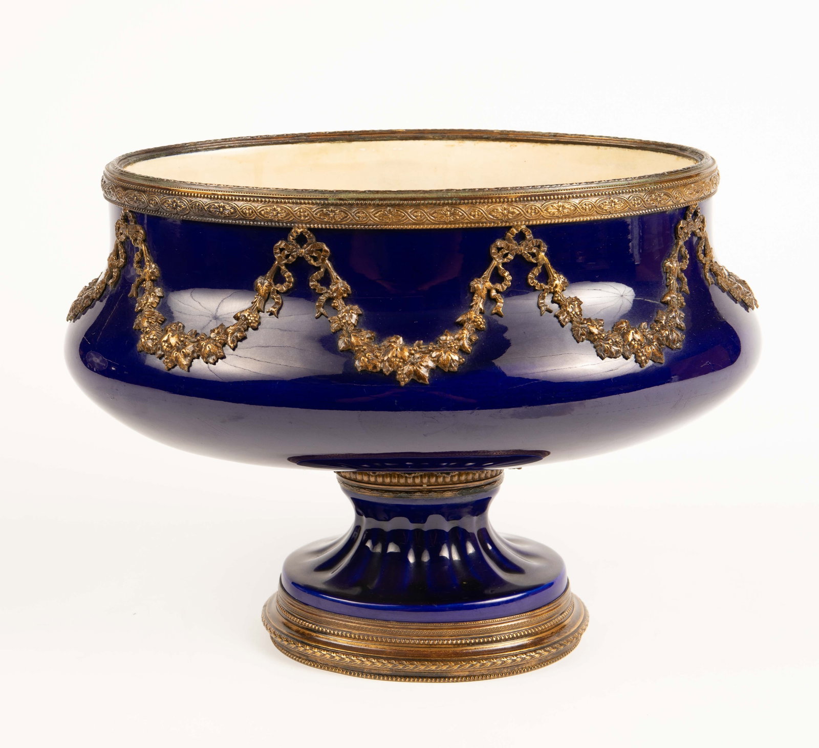MP Sevres Cobalt Blue Empire Style Planter Vase.: It is a cobalt blue footed planter with bronze mounts and ormolu garlands. The interior displays some crazing and discoloration, but overall, the piece looks to be in good condition. It measures 13 1/