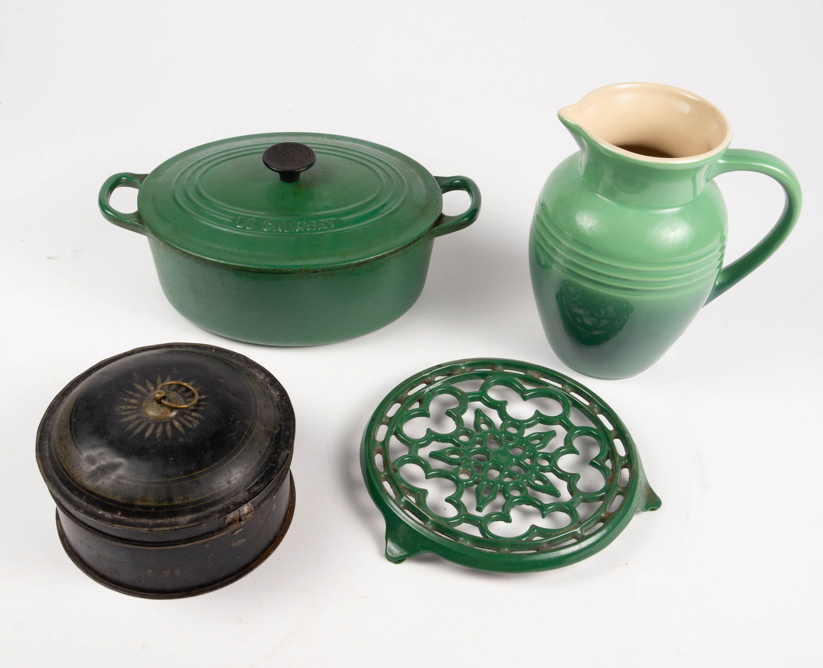 Lot: Le Creuset Green Items, Round Tea Tin.: The three Le Creuset items are: a ceramic water pitcher, a cast iron casserole pot with lid, and a Cousances cast iron trivet. The round black tin has dividers for different teas. Overall, the lot is
