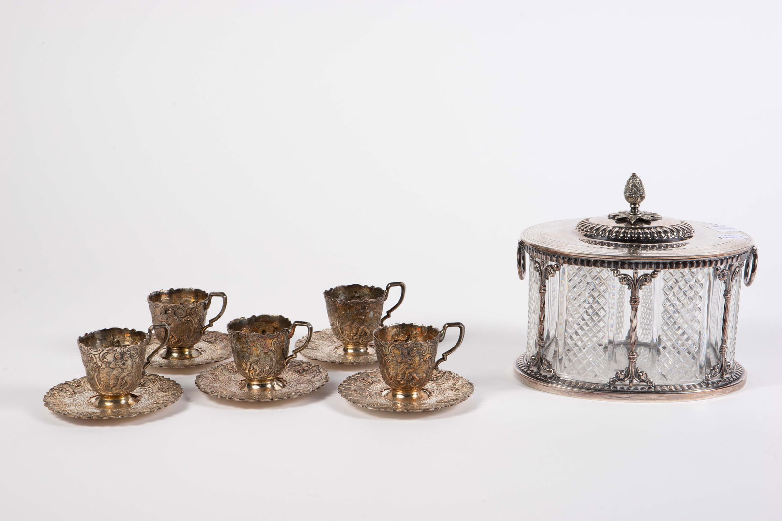 Lot: Silverplate Biscuit Barrel, Demitasse Set.: This lot includes a possibly Pairpoint silverplate and crystal biscuit barrel (has some small chips), and a set of 5 repousse silverplated teacups (no inserts) and matching saucers. Overall, the lot l