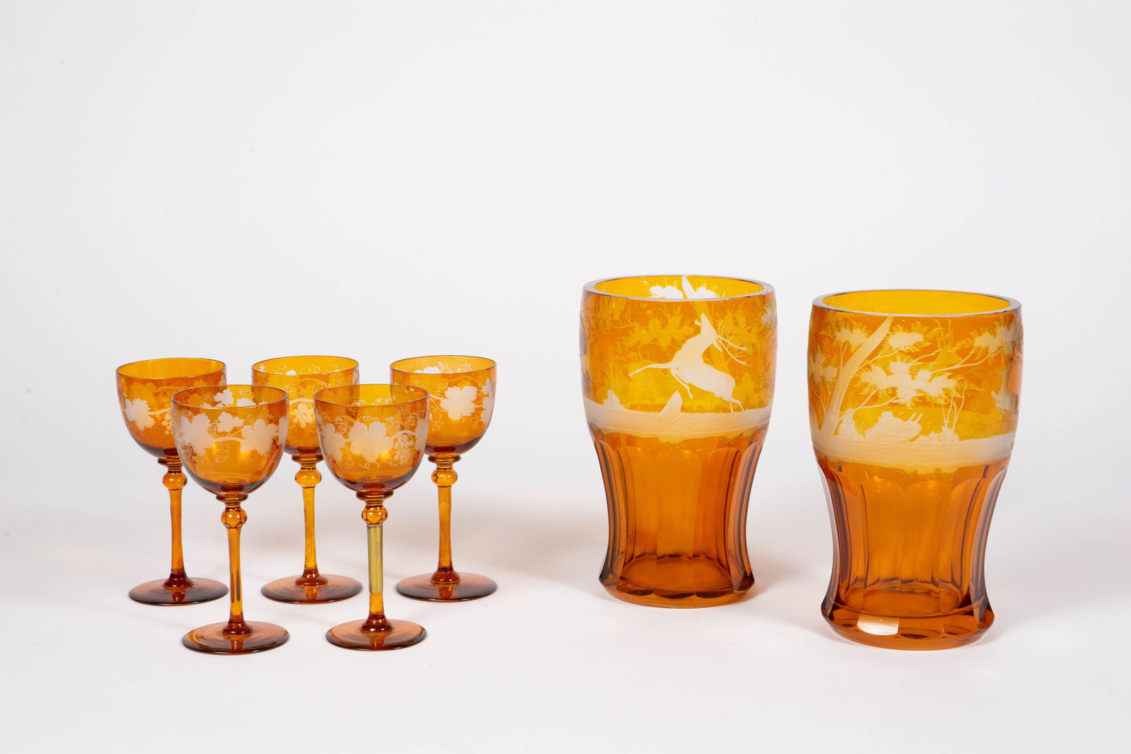 Lot: 7 Pcs. Bohemian Amber Glass w/ Etched Designs: The lot includes a pair of vases with deer forest scenes, and a set of 5 sherry glasses with grape leaf designs. Overall, the lot looks to be in good condition, with some minor fleabites and scratches