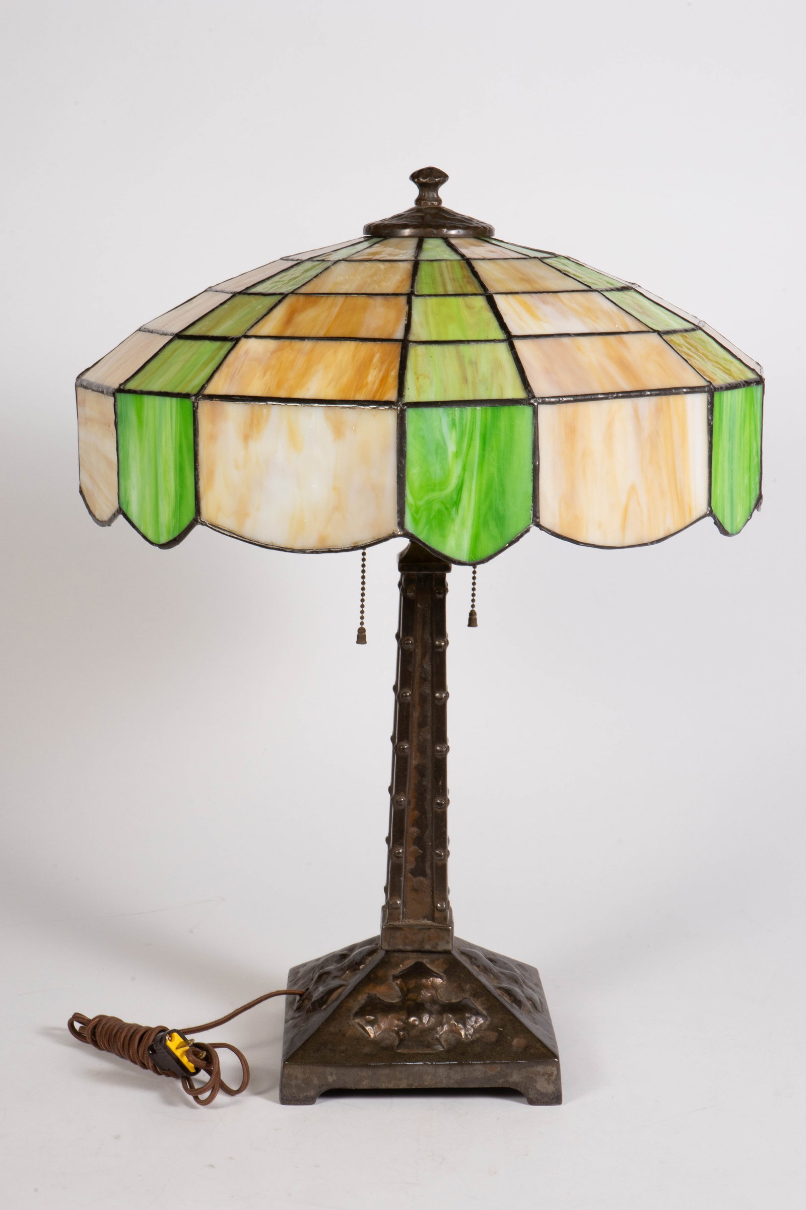 Arts & Crafts Leaded Glass Hammered Iron Lamp.: The lamp, circa 1910, features a hammered and tooled iron base, with nailhead and relief designs. The shade has green and beige slag glass, with a scalloped rim. The lamp measures 25 1/4" x 18 3/4" x 