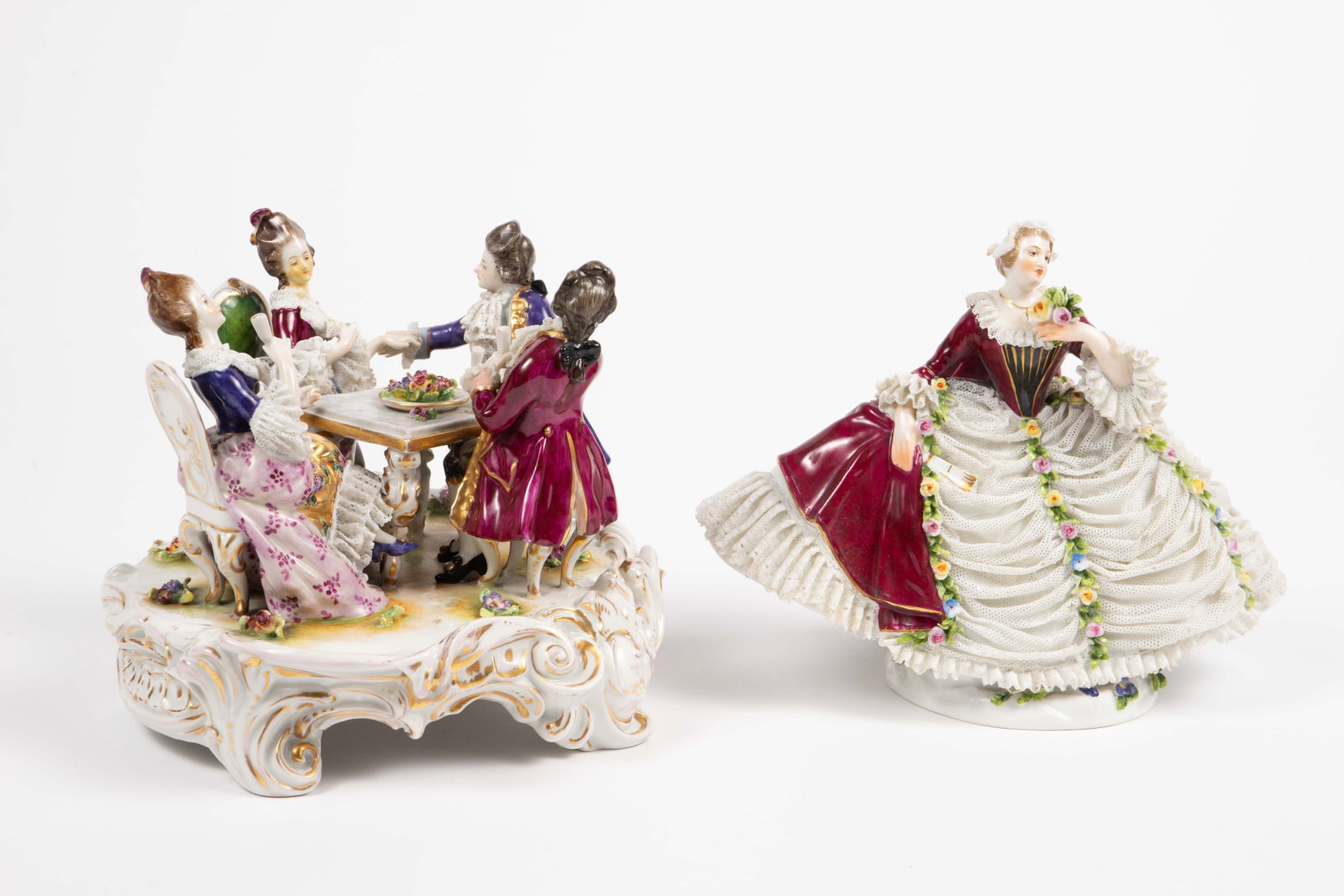 Lot: 2 Dresden Style Figures- Woman & Dining Group: The figural group depicts two couples seated at a table drinking wine. One of the women is missing her wine glass, and has discoloration to her face. The underside of the figure has a Volkstedt or Vol