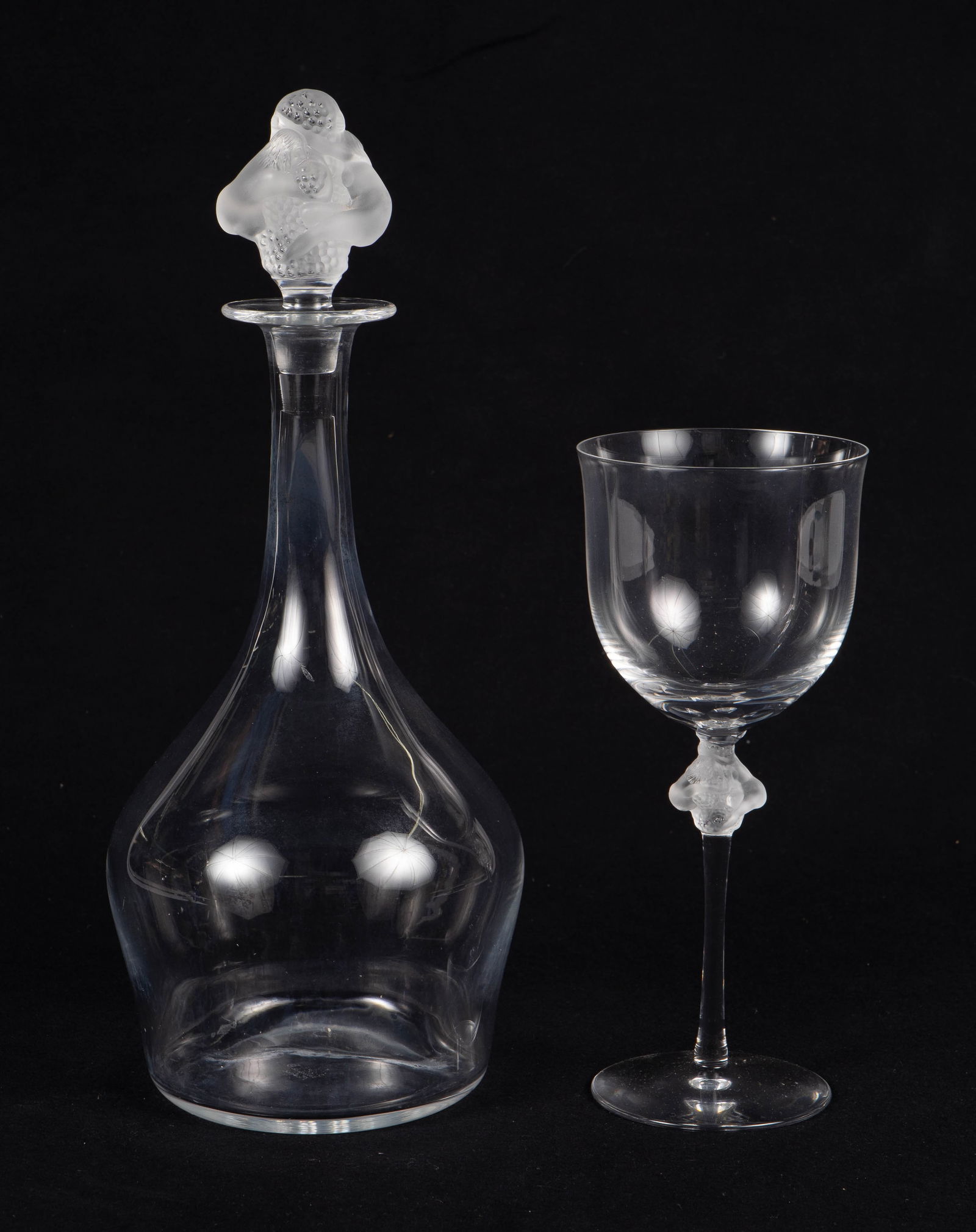 Lalique France "Roxane" Decanter and 12 Goblet Set: This lot includes a decanter and 12 matching wine glasses. The decanter's stopper resembles a pair of nudes with grape clusters. The knobs on the wine glasses match the stopper. Three of the goblets h