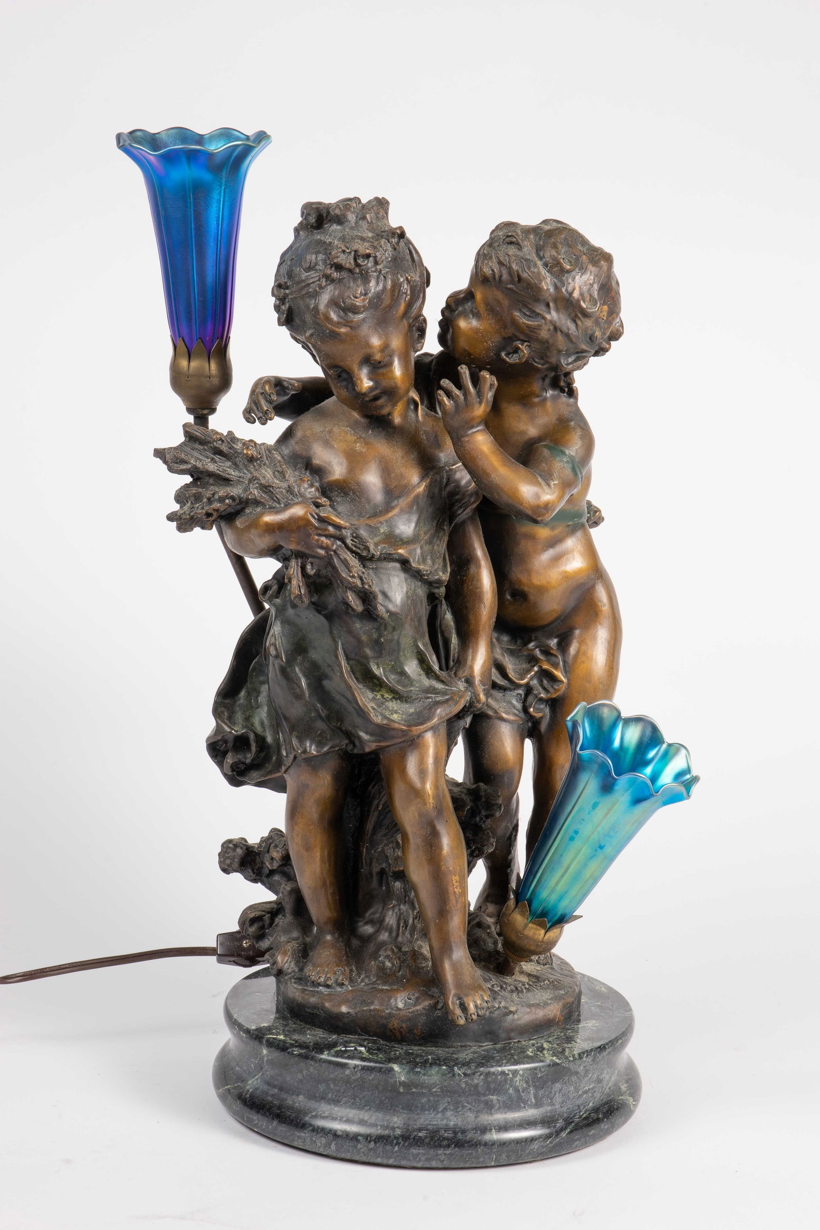 After Moreau Whispering Children Bronze Lamp.: The bronze sculpture after Auguste Moreau depicts a boy whispering into a girl's ear. The piece has two lights inside of blue iridescent floral shades, and a round green marble base. The bronze is sig