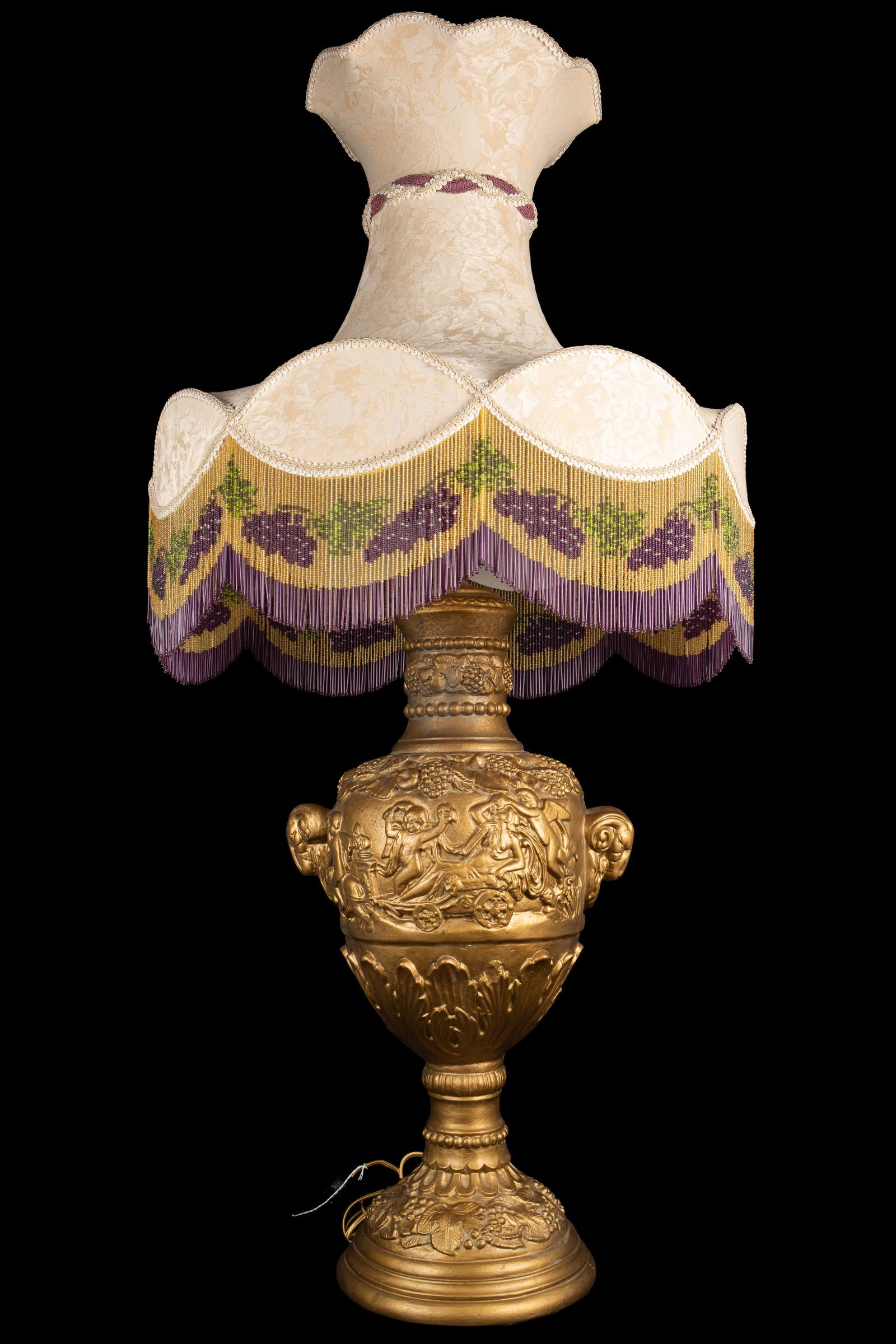 Repousse Style Lamp w/ Beaded Fringe.: The large lamp looks to be made of a plaster-like material, painted over with gold gilt. The lamp base features ram heads, cherubs, grapes, leaves, and more. The large shade has floral silk fabric and