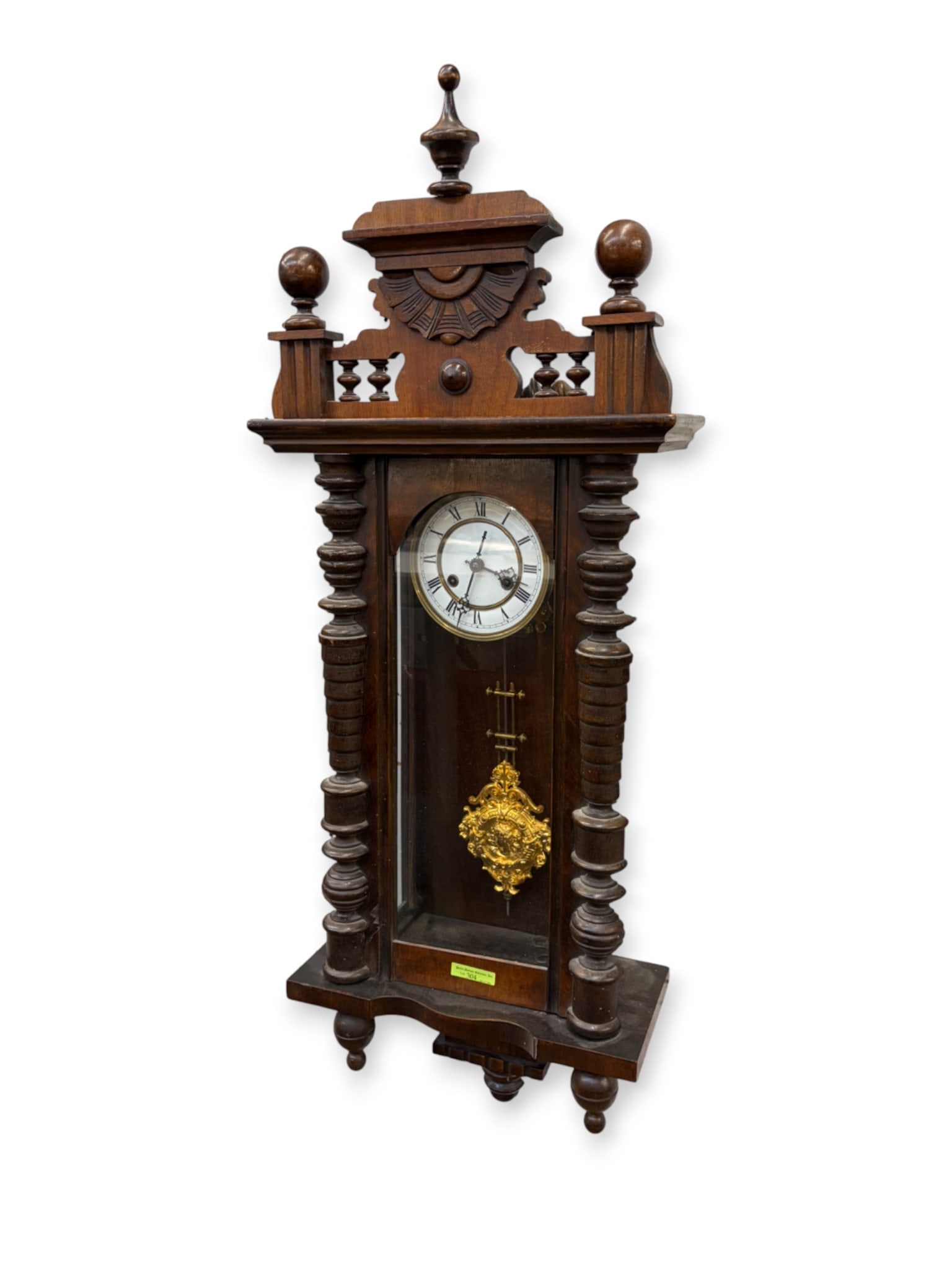 Antique Carved Wall Clock w/ Ornate Gilt Pendulum.: The antique time and strike wall clock features turned wood columns and finials, and a carved fan design decoration on the pediment. The porcelain face is unbranded, and the mechanism is not visible.