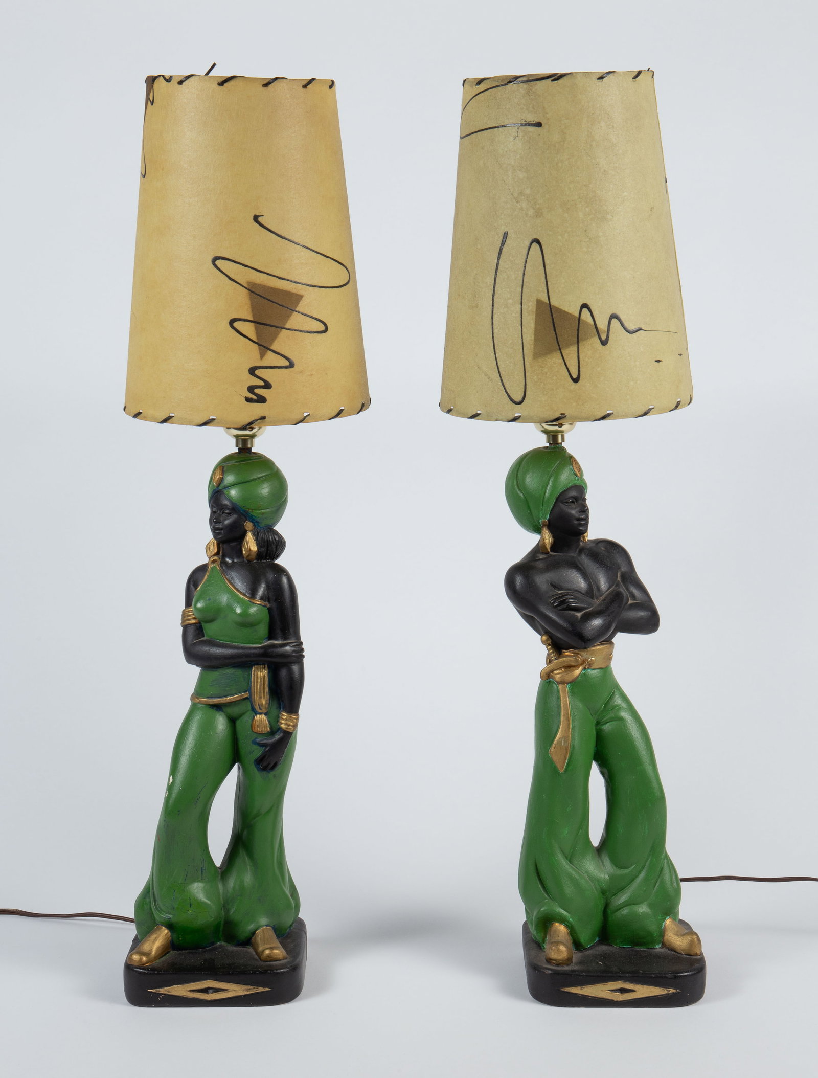 Pair MCM Blackamoor Nubian Genie Lamps.: The pair of chalkware lamps feature figural posts and abstract Mid Century shades. One lamp depicts a woman, and the other a man, both is green matching outfits. They measure 25 3/4" tall and 7 1/8" i