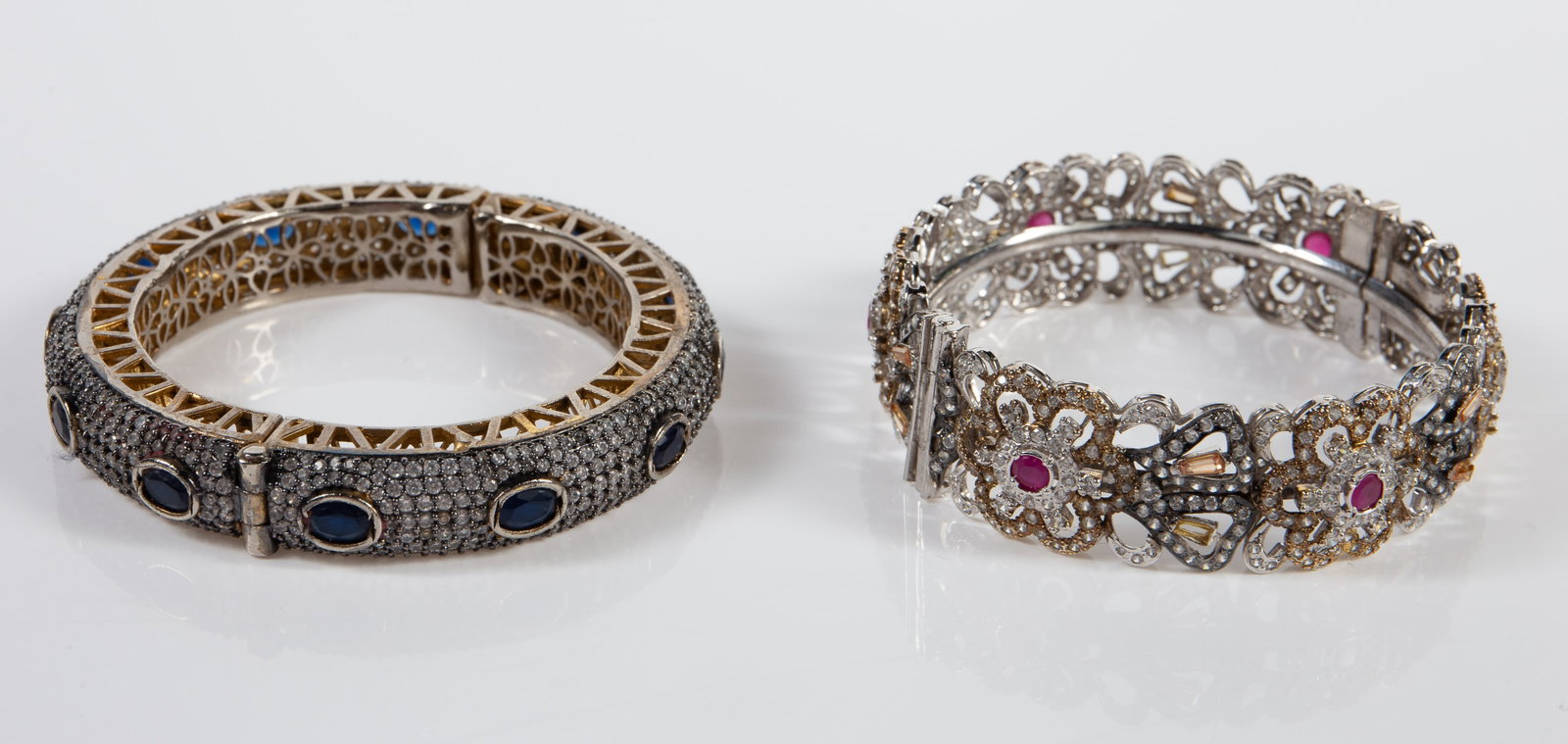 2 India Cuff Bracelets - CZ, Sapphire, Ruby.: The lot consists of 2 vintage India made bracelets with stones. One piece features pave cubic zirconia and oval blue sapphires. It measures 1/2" wide. The next piece has pierced flower decorations wit