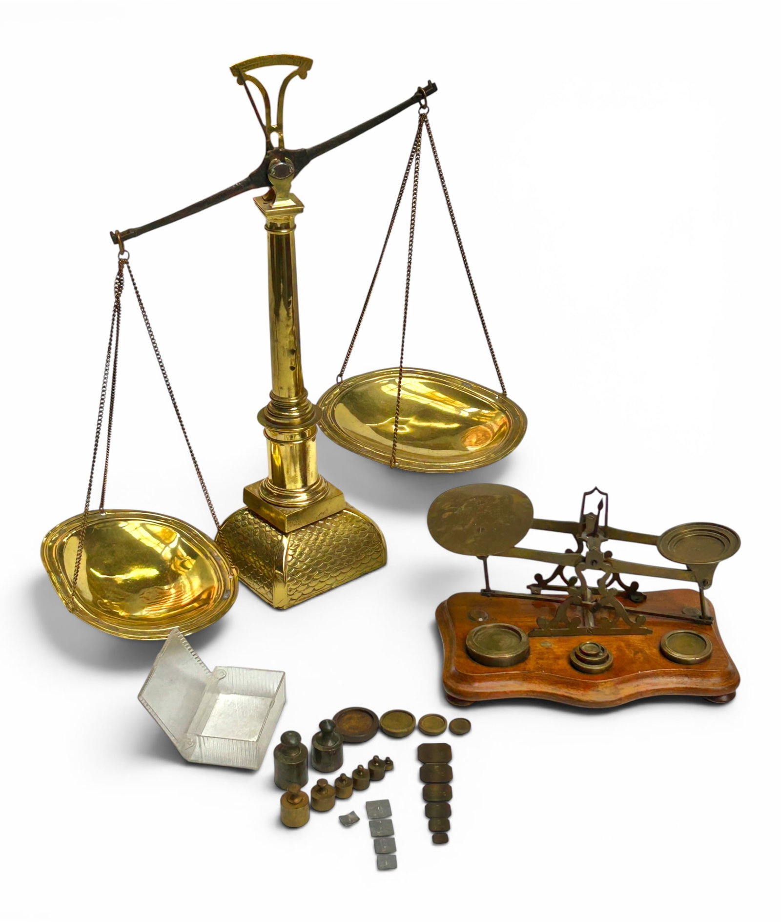 2 Vintage Brass Balance Scales, inc. Mitchell's.: The lot contains 2 brass balance scales. The largest one has two hanging shallow bowls and a fish scale pattern on the case. It has many various stamps near the central post. It measures 18 3/4" tall,