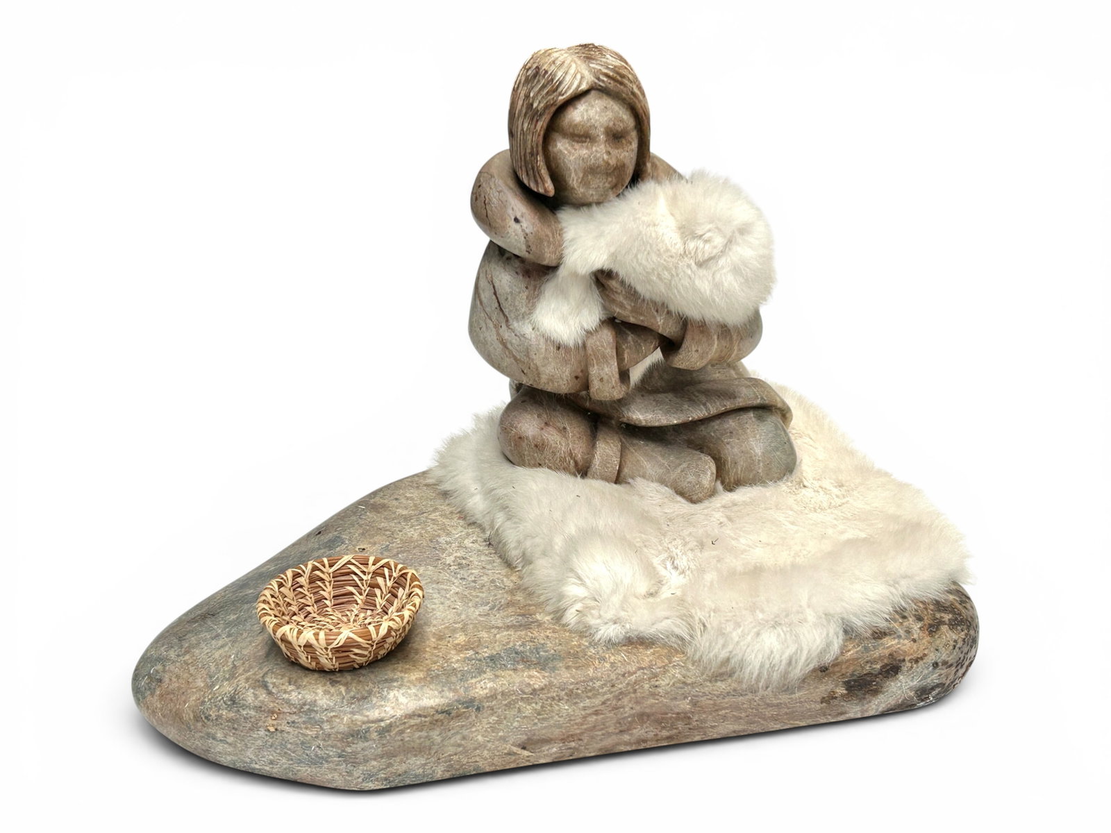 Inuit Carved Stone Figural Sculpture, Gene Drake.: The unique carved soapstone sculpture depicts a woman with short straight hair on a sheep skin rug holding a sheepskin bundle with presumably a baby in it. There is a woven basket sitting on the floor