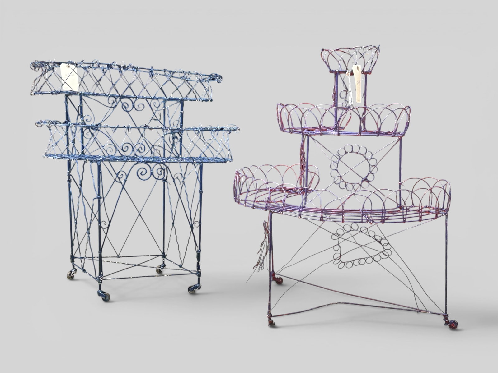 2 Victorian Style Wire Plant Stands Inc. Demilune.: The lot contains 2 painted wire plant stands on casters. There is a blue 2-tier piece, and a purple 3-tier demilune piece. The blue one measures 33" tall, 33 1/4" wide, and 17 3/4" deep. The demilune 