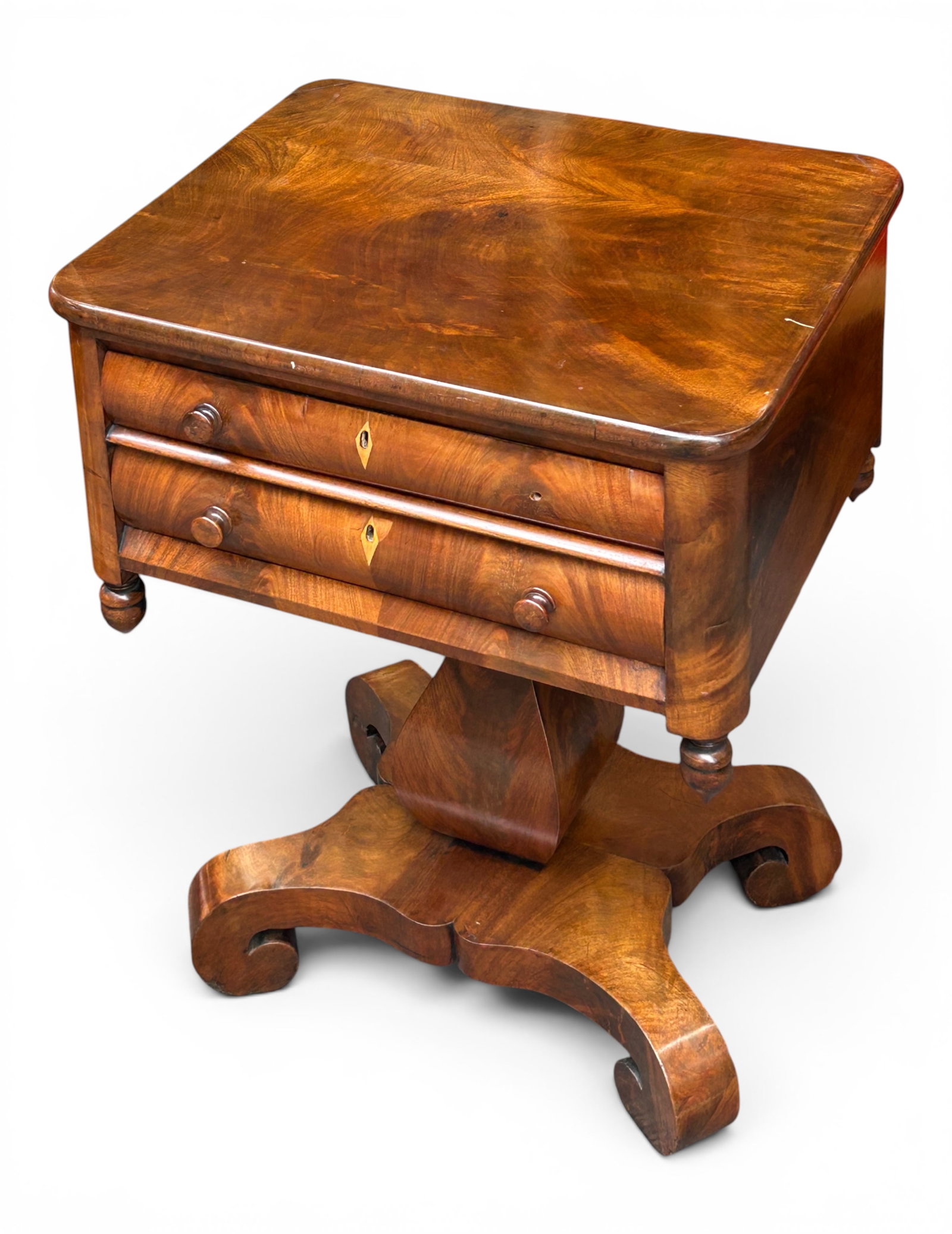 American Empire Mahogany Sewing Table.: This flame mahogany sewing table or stand has two drawers with wooden knobs, diamond-shaped escutcheons, finials on each corner, and a pedestal base. There are some repair spots and minor scratches.