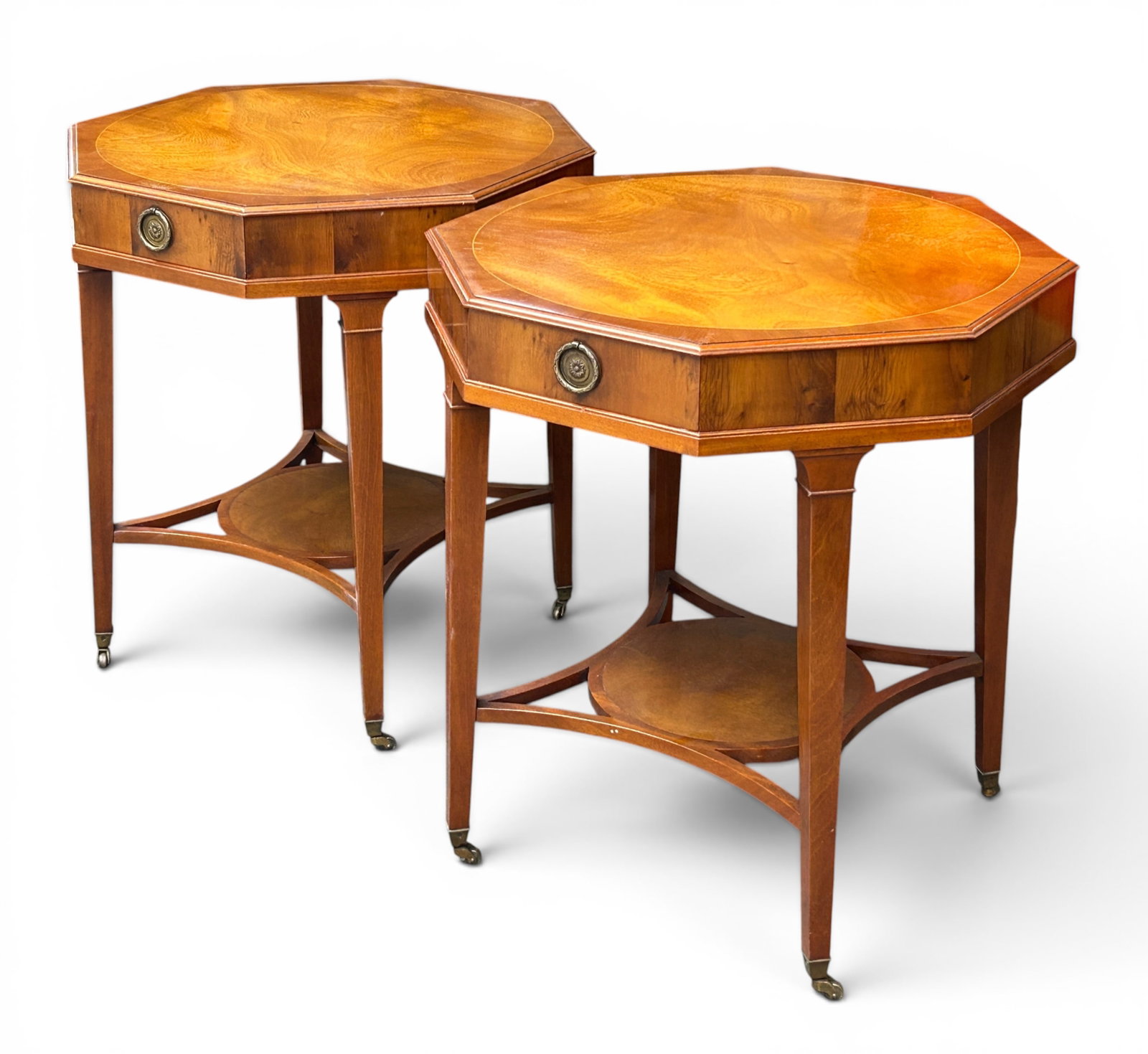 Pair of Inlaid Octagonal Mahogany Tables.: The tables each have an inlaid circular top, a single drawer, a brass pull handle, a lower tier shelf, and brass casters. Both are in good condition, with some light wear. Each one measures 24" wide,