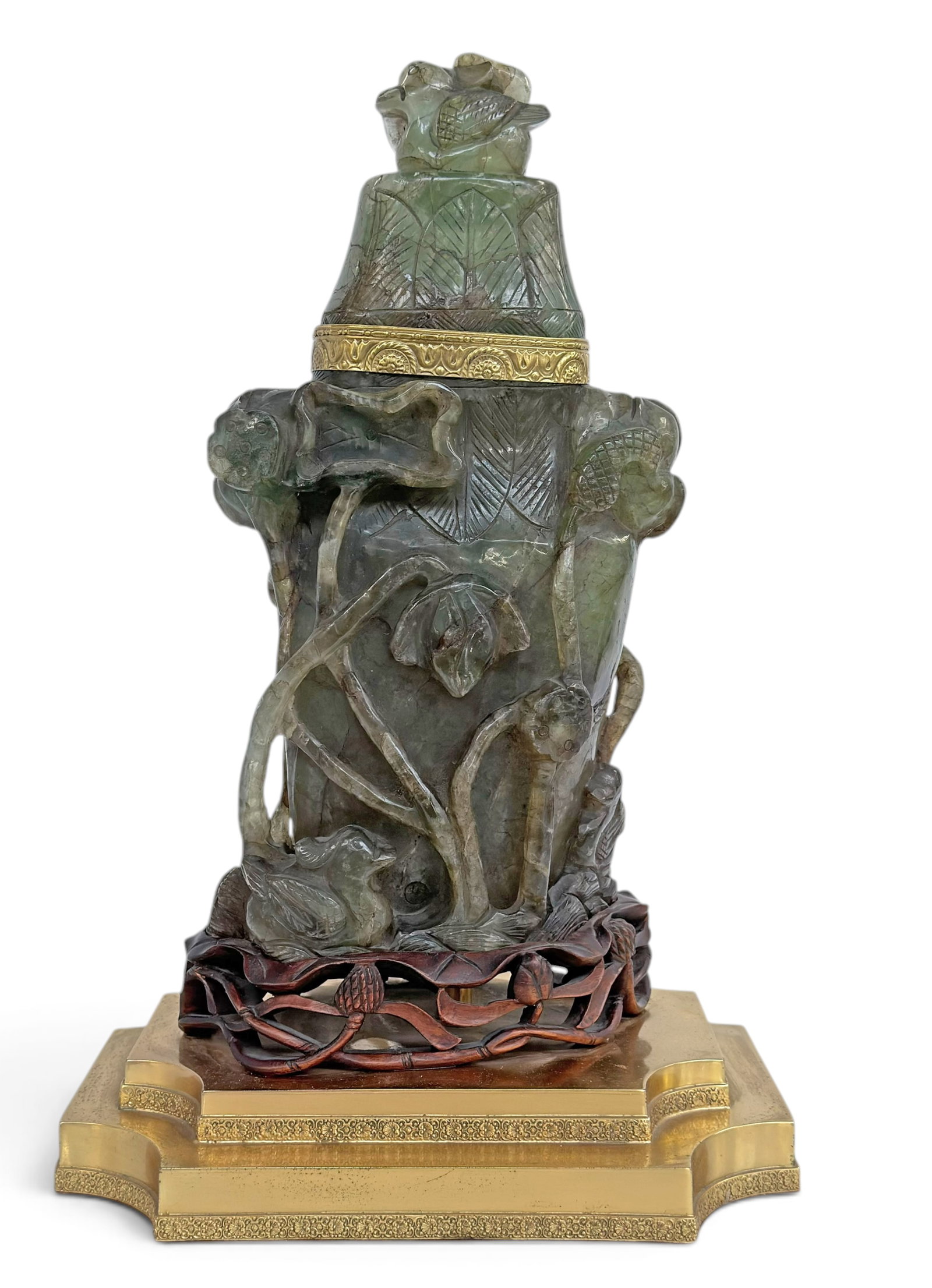 Large Chinese Carved Green Quartz Lidded Urn.: The piece is composed of a tiered brass base on a carved wood base, supporting the carved stone urn. The lid has a duck pediment, and the item is adorned with flowers and foliage relief. It measures 1