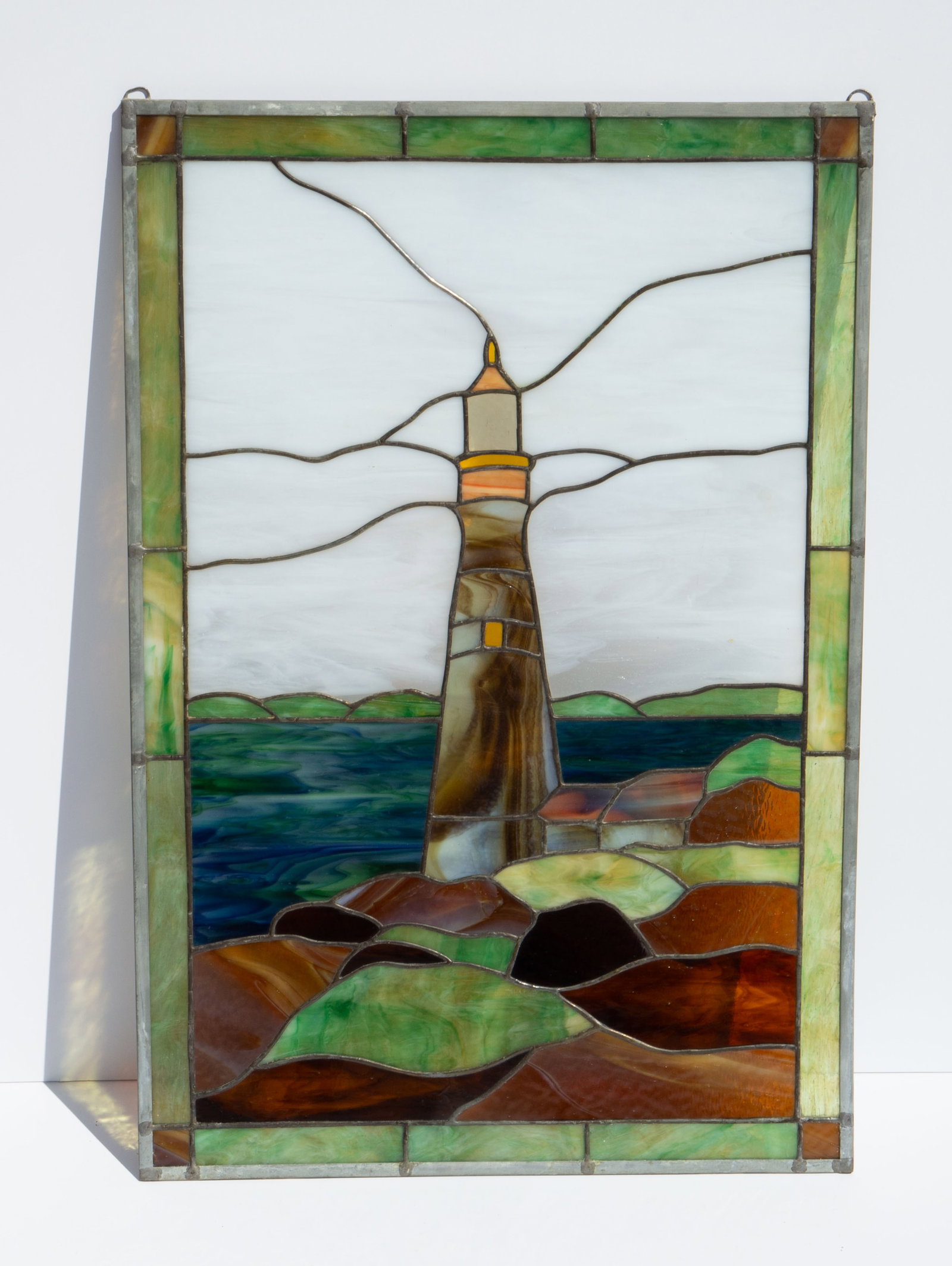 Leaded Lighthouse Scene Slag Stained Glass Window.: The decorative window features a coastal landscape with a lighthouse. The metal frame measures 30" x 20". There are some scratches and stains on the piece. Otherwise, the item is in overall good