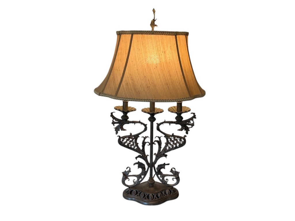 Ornate Bronze Table Lamp with Fabric Shade, 36"H: Bronze table lamp featuring an intricately detailed base with scrollwork and leaf motifs. The lamp is fitted with a light tan, pleated fabric shade adorned with decorative trim. Three socket arms crea