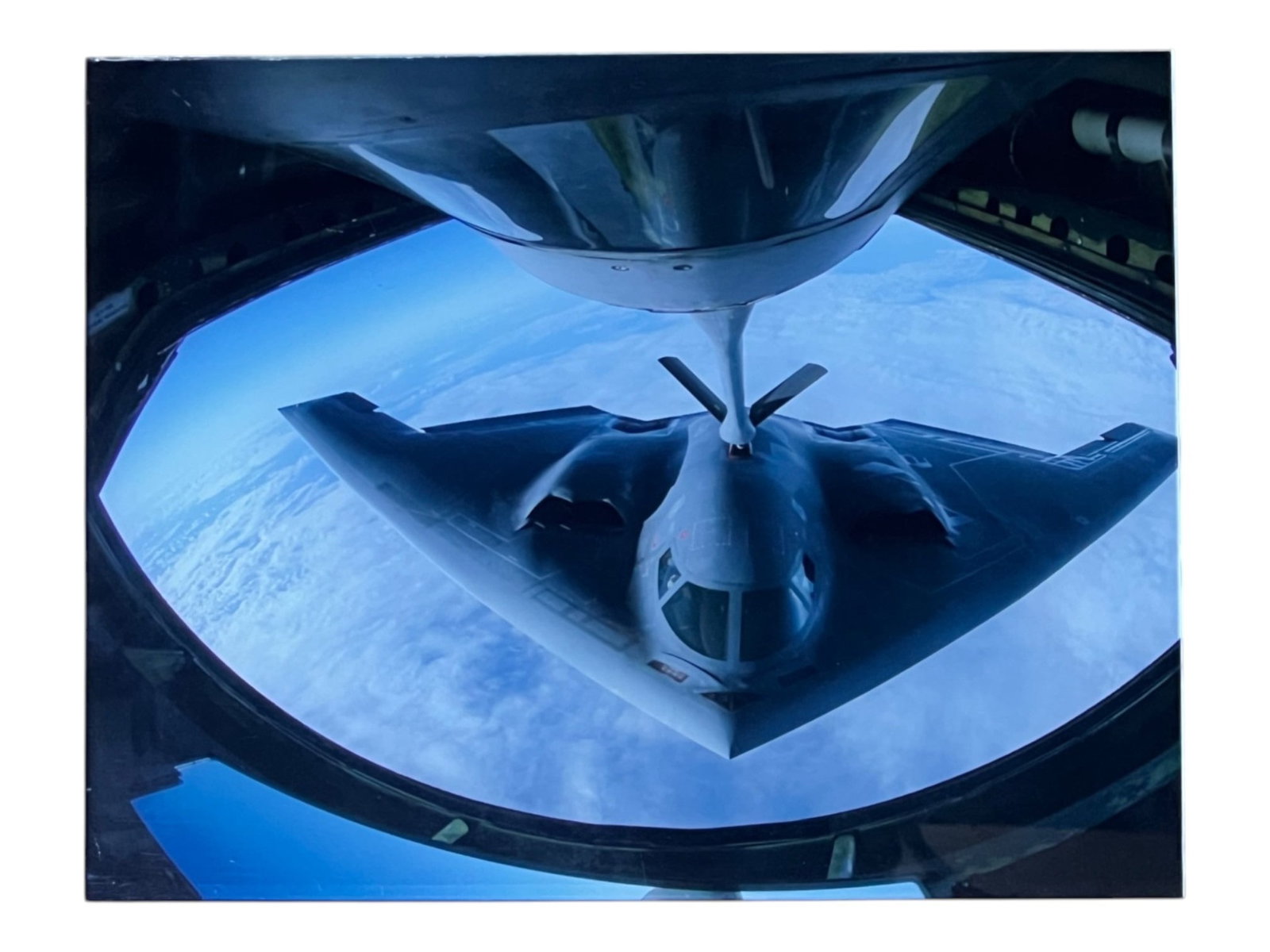 Contemporary Aerial Photograph, B-2 Spirit Stealth Bomber in Mid-Air Refueling, Plexiglass Mounted,: A striking aerial photograph capturing a B-2 Spirit stealth bomber during mid-air refueling, viewed from the boom operator's station of a tanker aircraft above. The bomber's distinctive flying-wing
