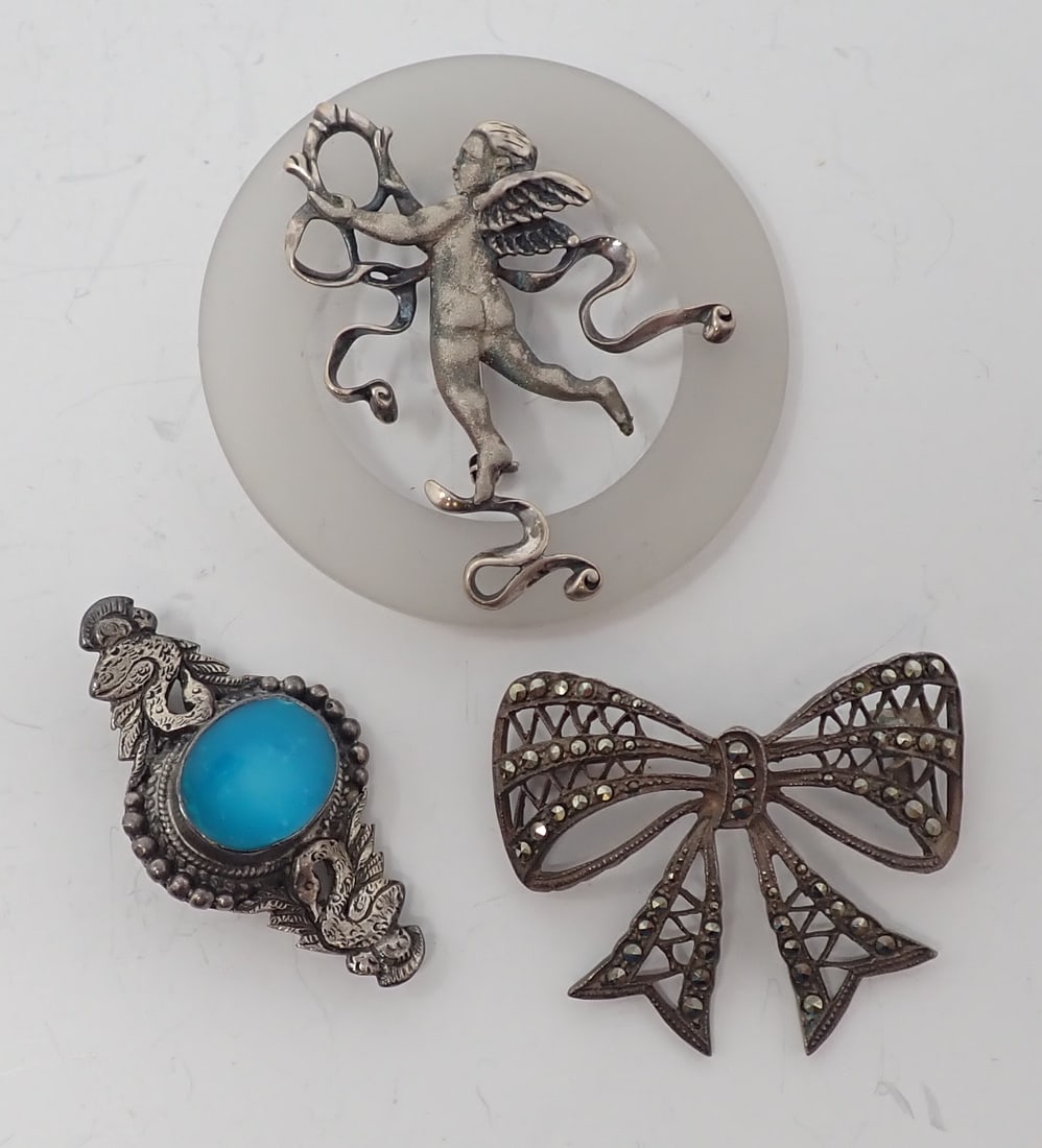 3 Sterling Brooches: Includes a form of a puti or cupid over a frosted plastic ring; a double swan brooch with a center bezel set oval hemimorphite; and an unmarked bow shaped brooch with marcasites. 1.20 ozt total.