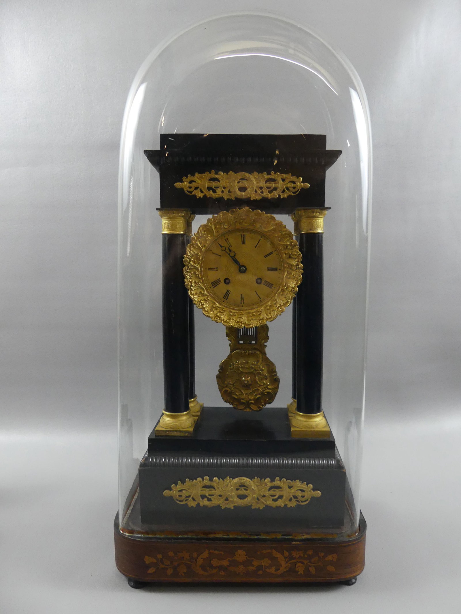 FRENCH PORTICO CLOCK UNDER DOME: Antique ebony wood French portico clock with ormolu mounts and fancy brass pendulum. Unmarked brass movement. Clock is 18 in high x 9.5 x 5.5 deep. Comes with fine glass Victorian dome with inlaid ros