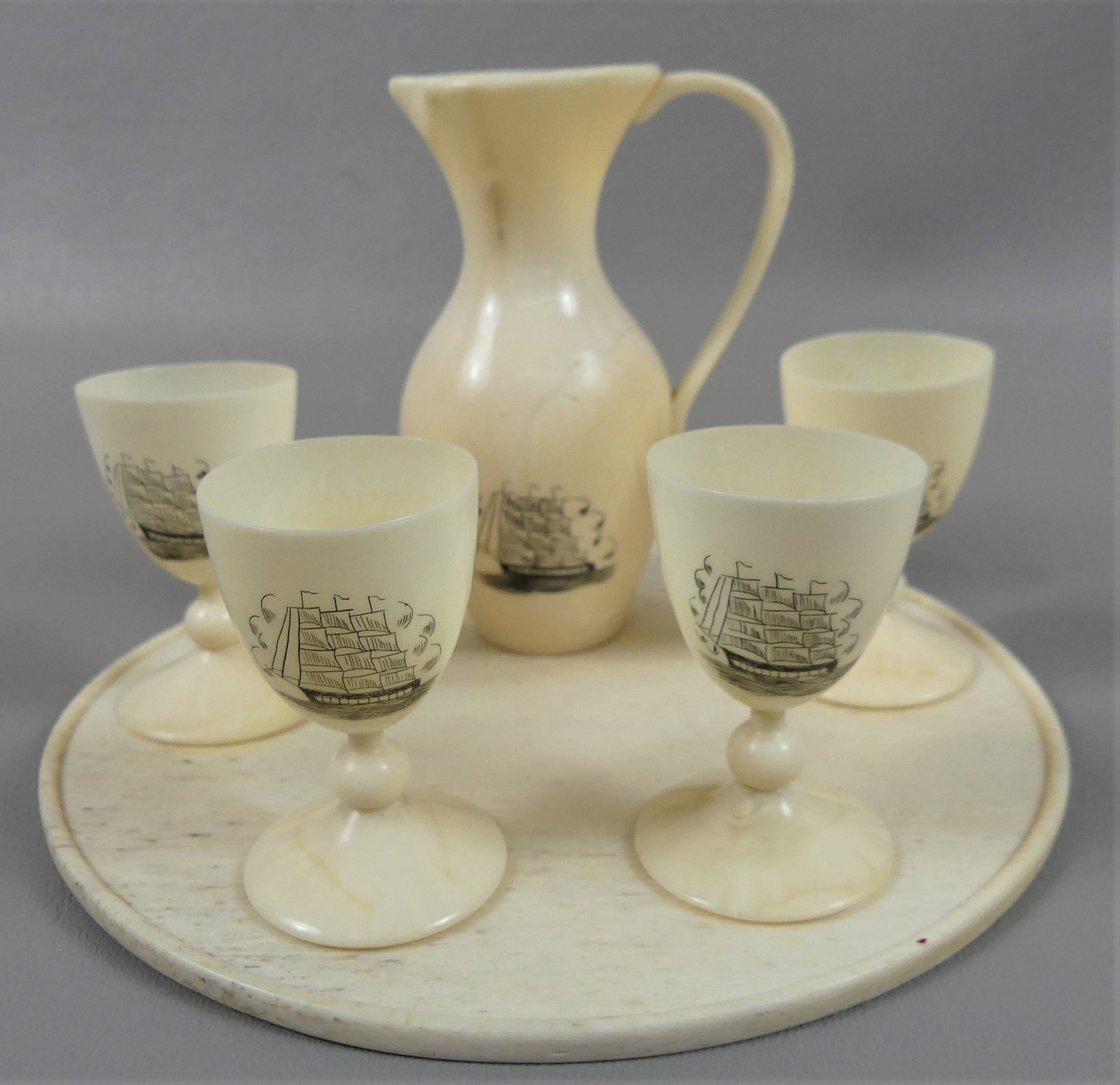 SAILOR MADE WHALE BONE CORDIAL SET: Fine 6 piece carved whale bone cordial set with scrimpshaw of ships. Consists of a 3.75 in pitcher; 4 footed cordial cups, 2 in; and 6 in round tray. VG condition, hairline in one cup, filled crack on