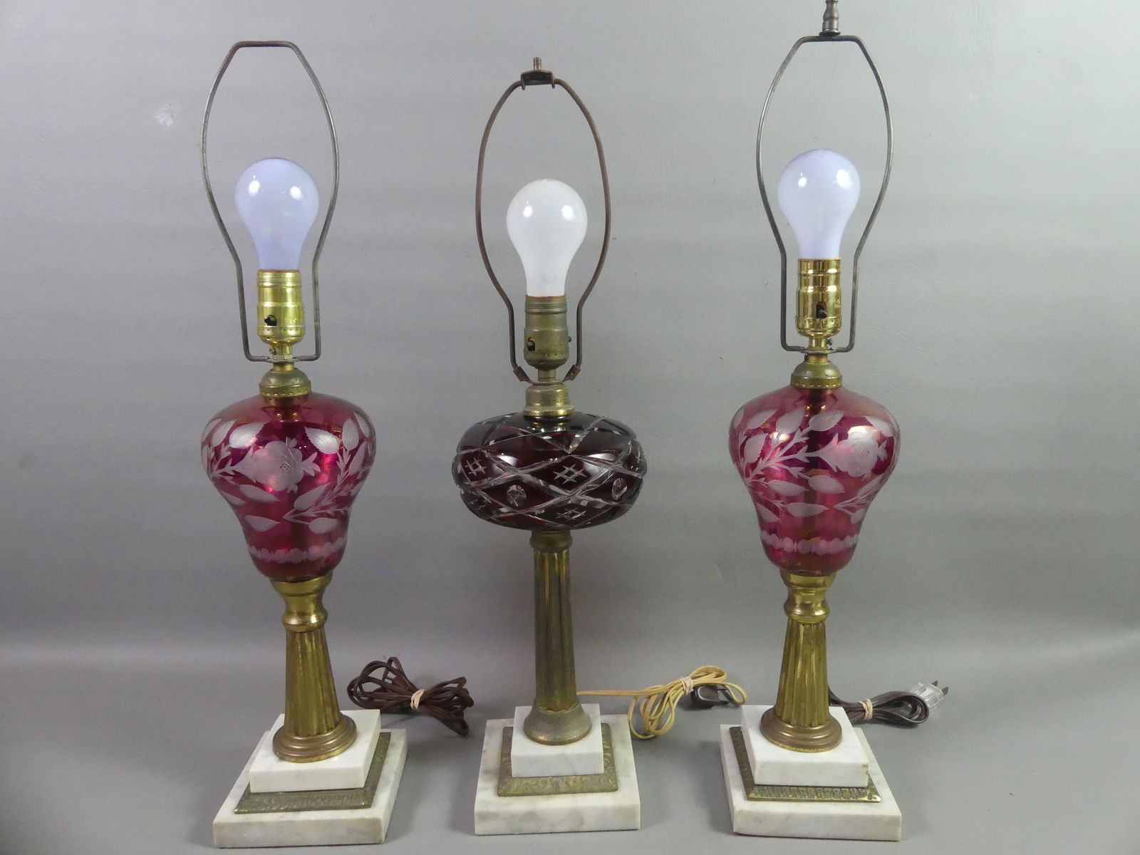 3 ANTIQUE CUT GLASS LAMPS: Lot of 3 antique lamps (electrified) with cut glass fonts - pair of cranberry cut to clear fonts with flowers, and ruby geometric design. They have white marble and brass bases. To 24 in tall.