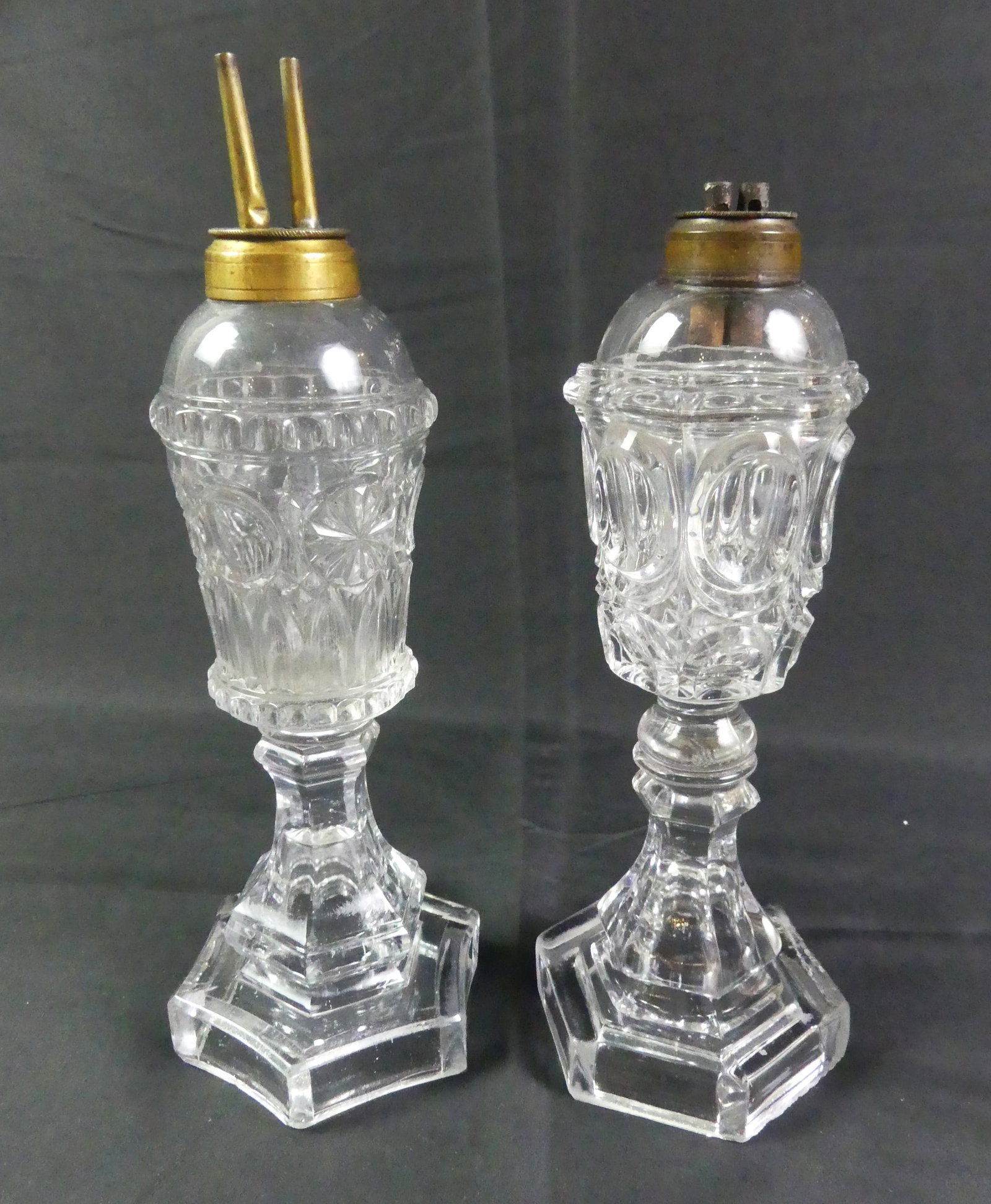 2 SANDWICH GLASS WHALE OIL LAMPS: Lot of 2 Sandwich flint glass whale oil lamps to 10.5 in tall with original brass double burners and hexagonal bases. Good condition, couple shallow flakes on underside of thumbprint lamp.
