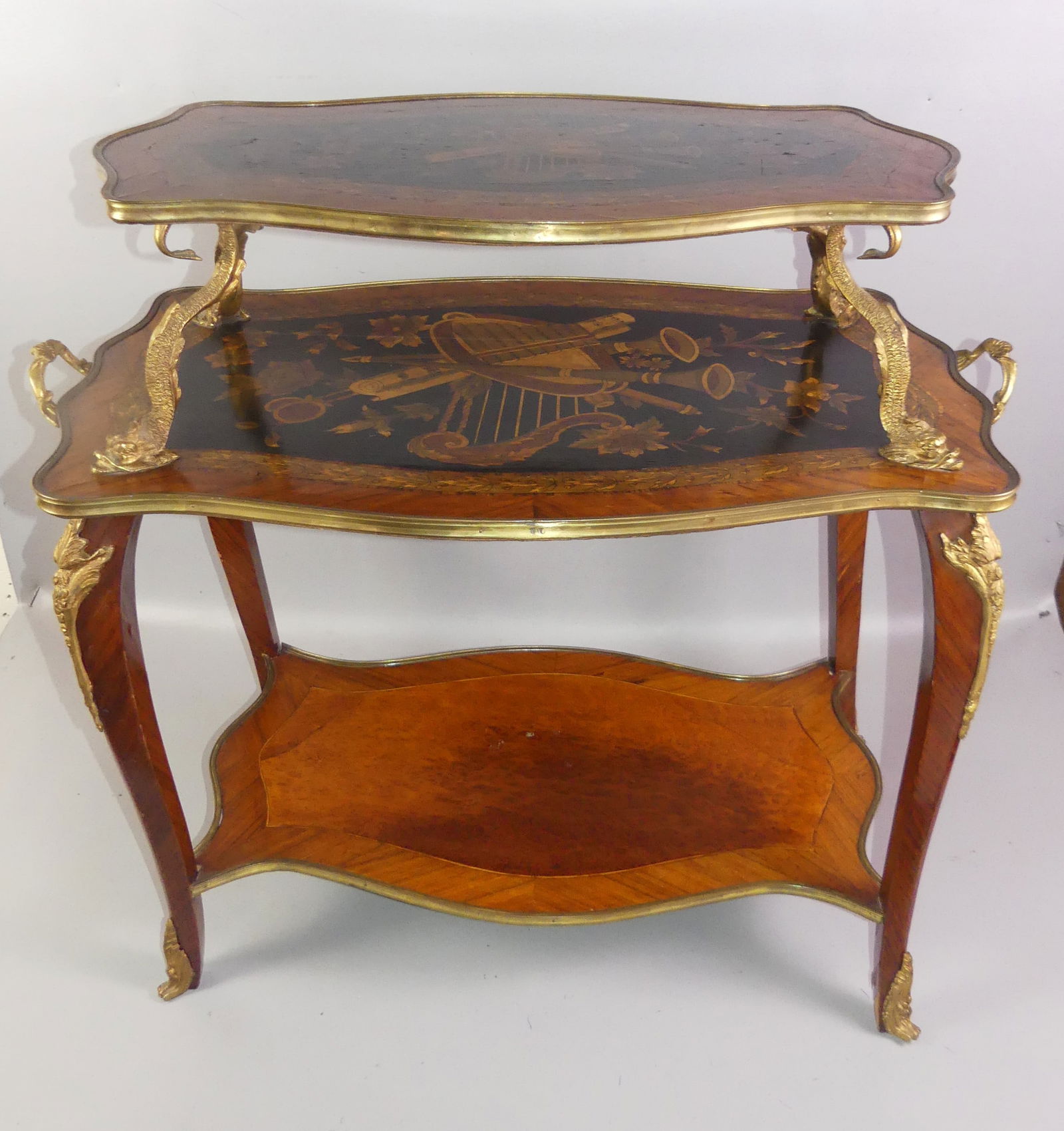 INLAID FRENCH 3 TIER TABLE: Old 3 tiered French Louis XIV satinwood table with 3 shelves, 2 having elaborate inlay of musical instruments, flowers, etc. Ormolu mounts, handles, and dolphin supports. 30 in high, 34 wide and 19.5