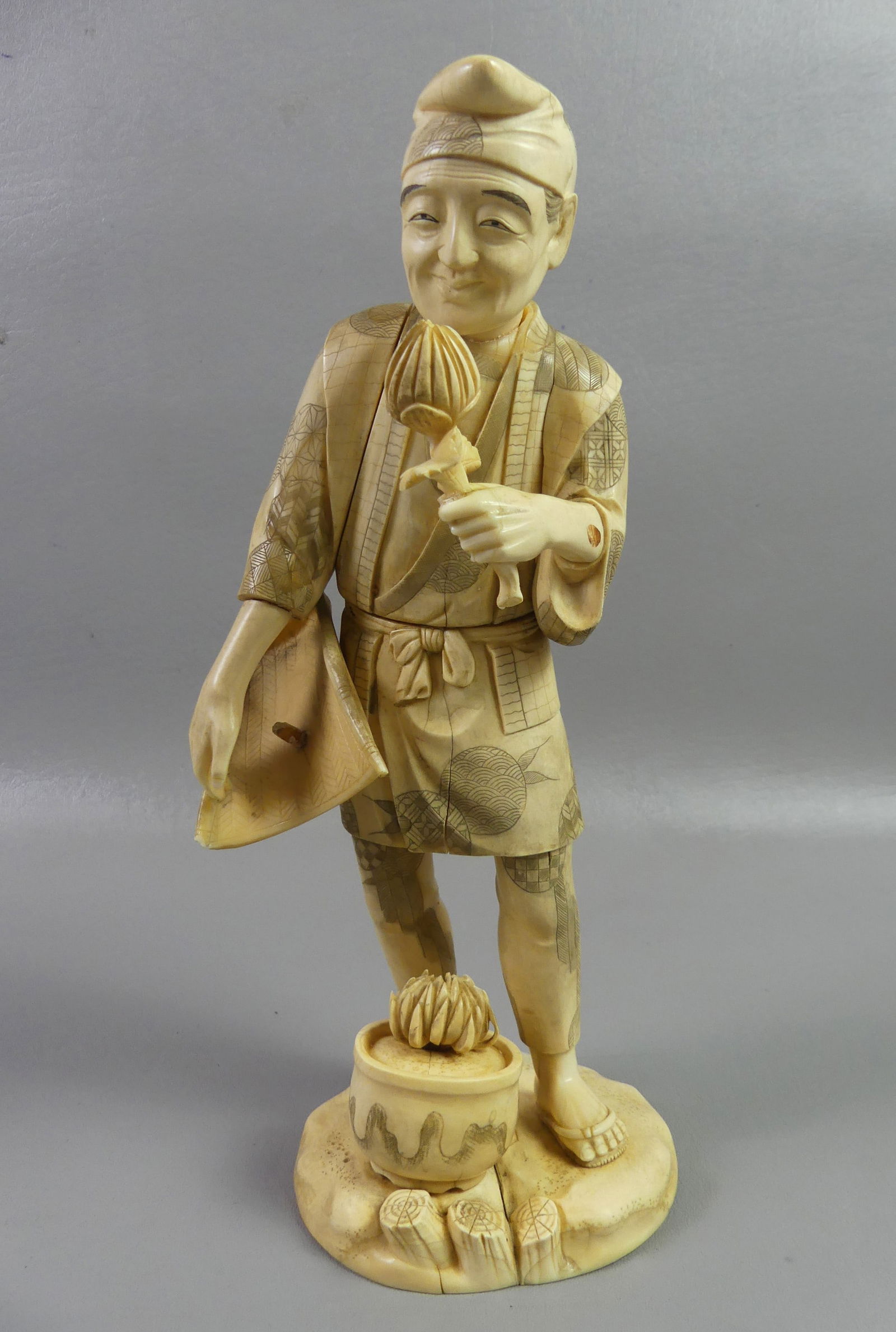 JAPANESE CARVED BONE MERCHANT: Japanese Meiji period carved figure of an Asian flower seller or merchant carrying basket and holding a flower with a flower pot at his feet. Signed on underside with Asian characters. 11.5 in tall,