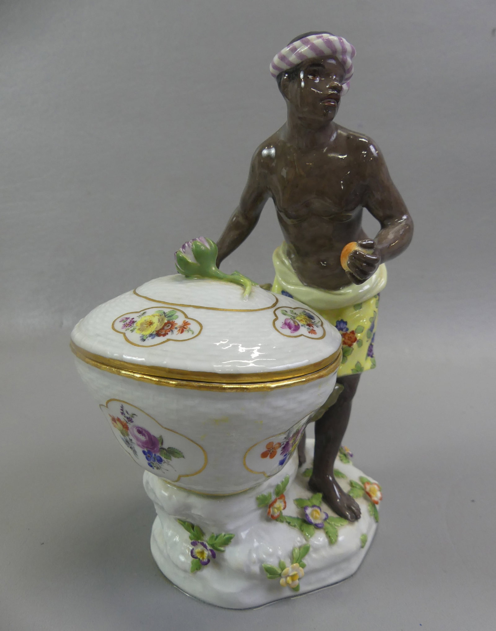 MEISSEN FIGURAL SWEETMEAT DISH - BLACKAMOOR: 18th century Meissen porcelain figural sweetmeat dish of a blackamoor with bandana and yellow skirt holding a covered bowl with scattered applied flowers. Has crossed swords underglaze mark and #336.