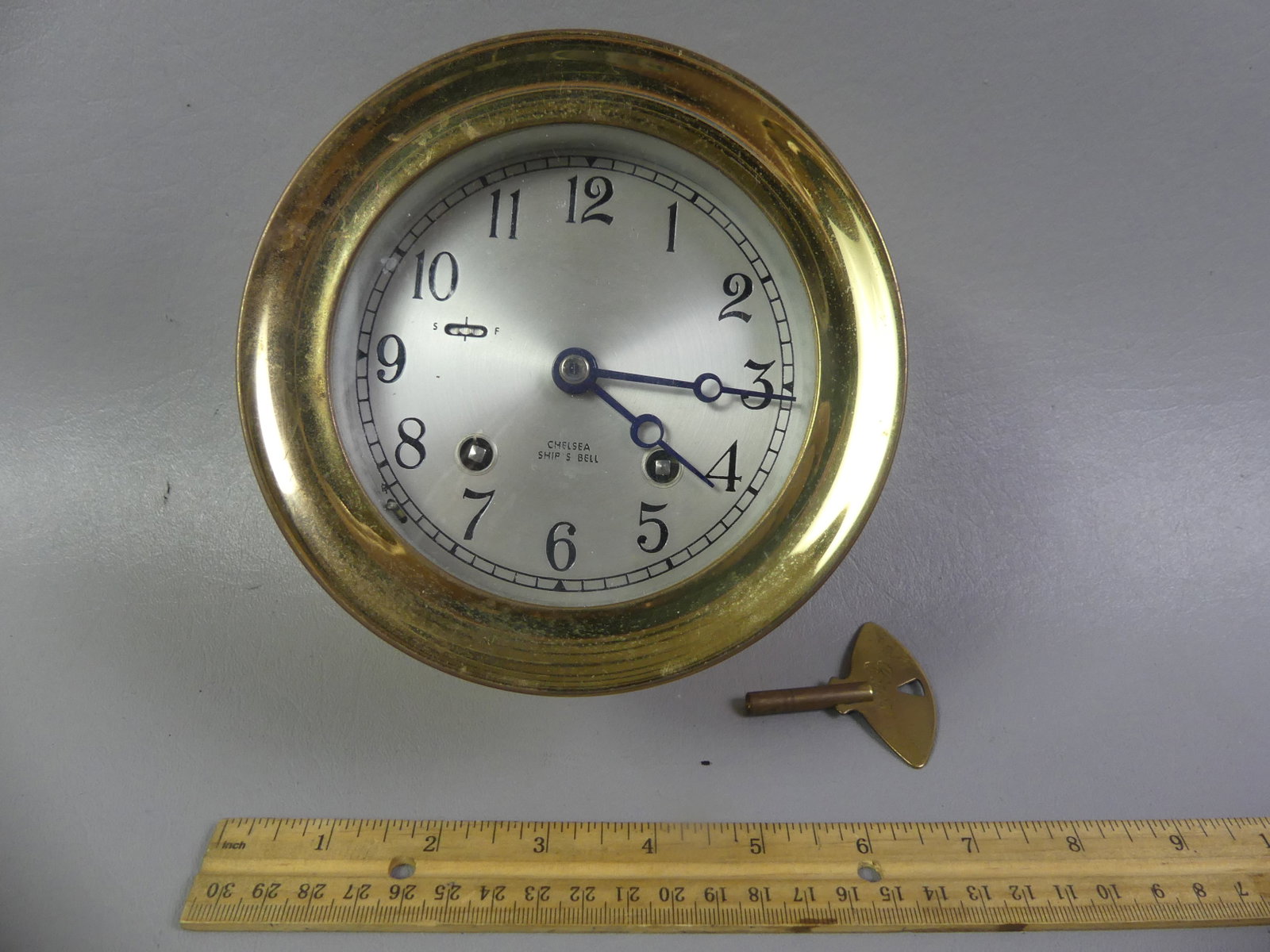 CHELSEA BRASS SHIP'S CLOCK: Old ship's bell wall clock in brass signed Chelsea, 4 in dial, 5.5 in diameter bezel. Currently working with key.