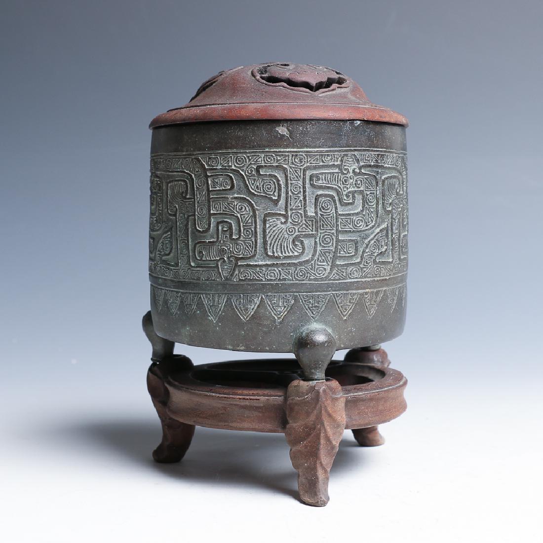 BRONZE INCENSE BRUNER WITH A STAND: Of cylindrical form supported on three short legs, scrolling designs and patterns band carved on the body, accompanied with a wooden lid with three ganoderma carving peiced through and a fitted wood t