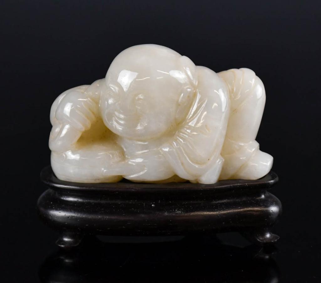 A Chinese Jade Carved Boy w/ Woodstand & Box Qing: Carved as a smiling boy dressed in a patterned robe with a drum, the stone of a celadon tone with russet inclusions. L: 6cm 清 白玉童子擺件連底