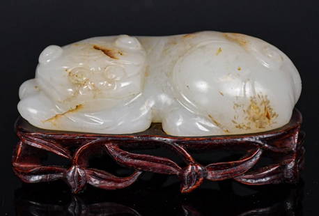 A White Jade Carved Beast W/ Std Qing: A beast carved from a white jade stone with russet speckles, in a prone position. With the beast's head turns to it's left. W: 4.5cm, L: 8.3cm 清 白玉雕瑞獸擺