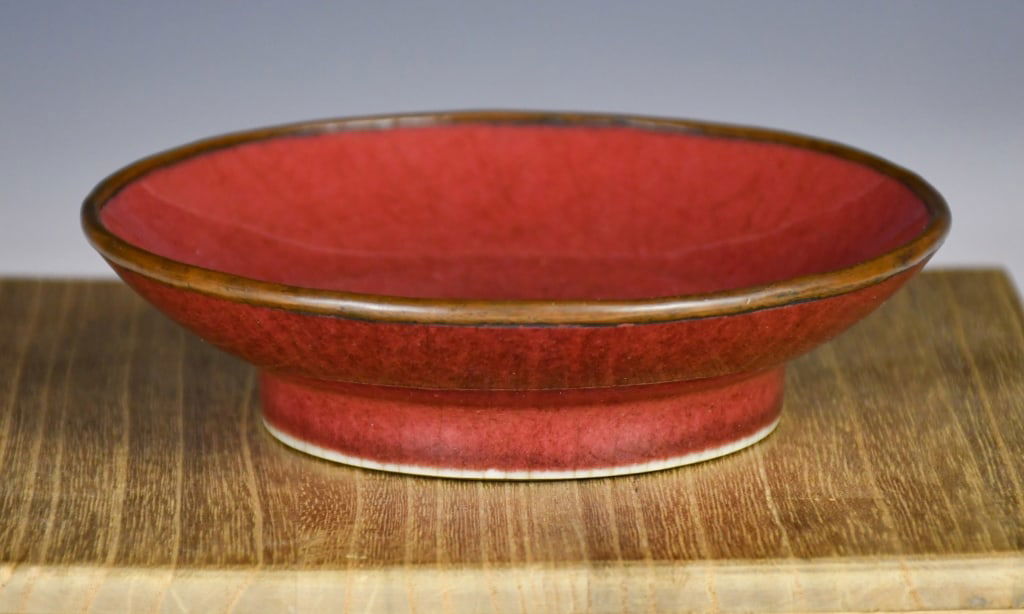 A Oxblood Glazed Dish Ming Dynasty: The shallow dish of rounded form, supported on a short foot, covered overall in a rich oxblood-red glaze stopping neatly above the foot to reveal the pale bottom. D:22.8cm 明 紅釉