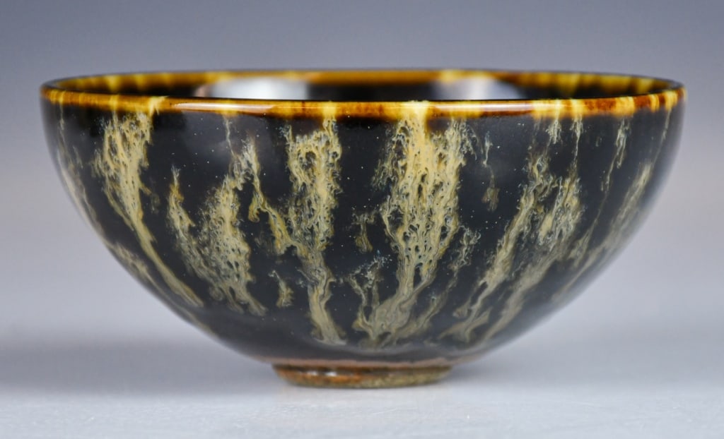 An Archaistic Jian-Kiln Tea Bowl: Modeled with deep U-shaped sides rising from a short foot, covered overall in a dark glaze suffused with irregular splashes, the rim highlighted with brown glaze. H: 5.2cm, D: 10.9cm 高古&