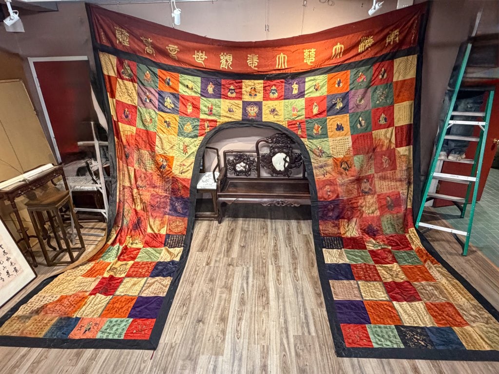 A Large Embroidered Canopy Qing: The silk canopy composed of polychrome patchwork panels, each embroidered with Buddhist figures, auspicious motifs and calligraphic inscriptions, bordered in dark silk and surmounted by a wide 