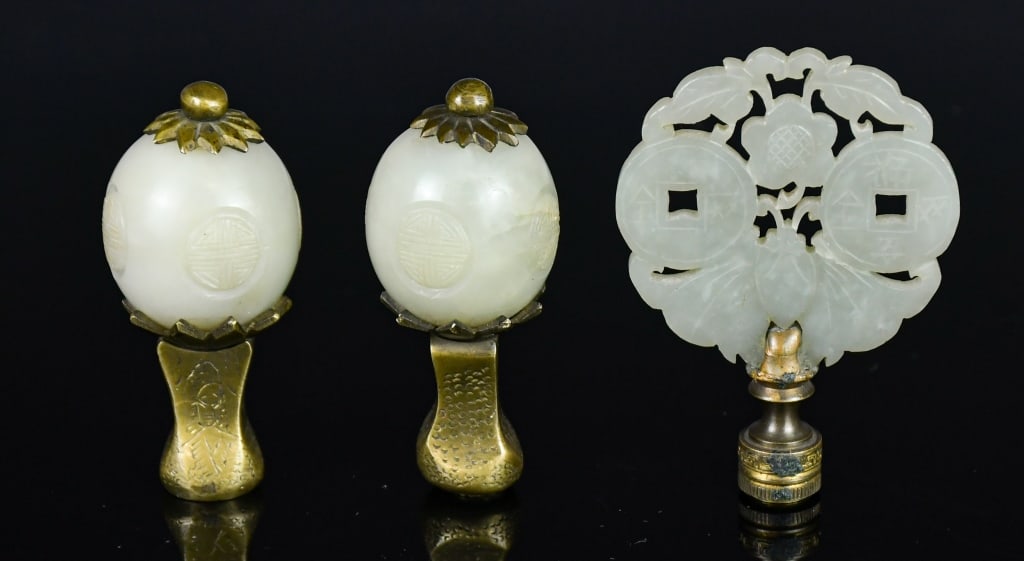 A Group of 3 Jade Lamp Finials 19thC: Comprising three jade finials, two of oval form carved in low relief and mounted within settings, each raised on a shaped base; the third of circular openwork form carved with two coin motifs, a