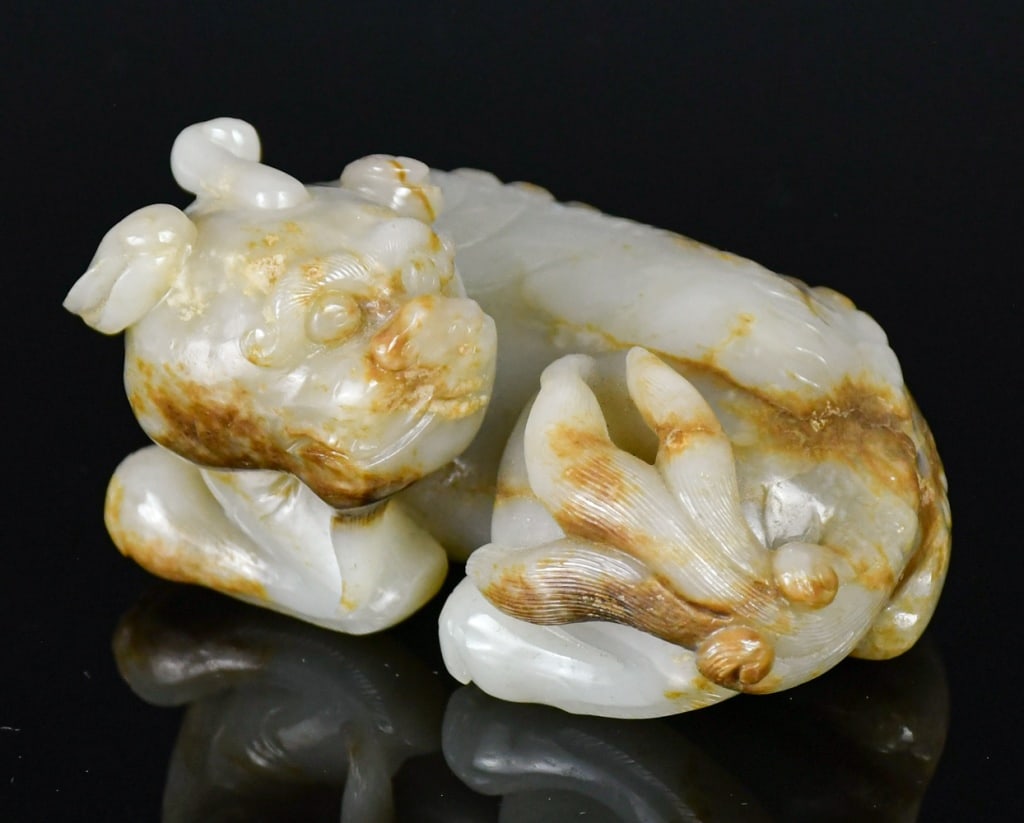 A Celadon Jade Carved Mythical Beast: Carved from a pale celadon jade stone with russet inclusions, depicted a mythical beast in a crouching position. 11.1x8.5cm 青玉瑞獸擺件 Click here for