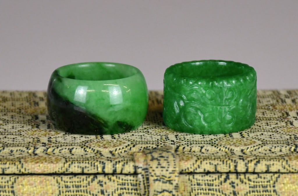Group of 2 Jadeite Rings w/Box: Comprising two jadeite rings of differing size and form, one carved to the exterior with decorative motifs, US size:8.5; the other of plain design, US size: 9.25. 