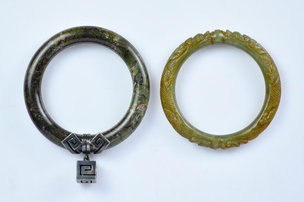 A Group of 2 Celadon Jade Carved Bangles Qing: Comprising two jade bangles, one of plain circular form, the stone of dark celadon tone suffused with reddish veining; the other carved in relief with two confronting dragons with a pearl, the 