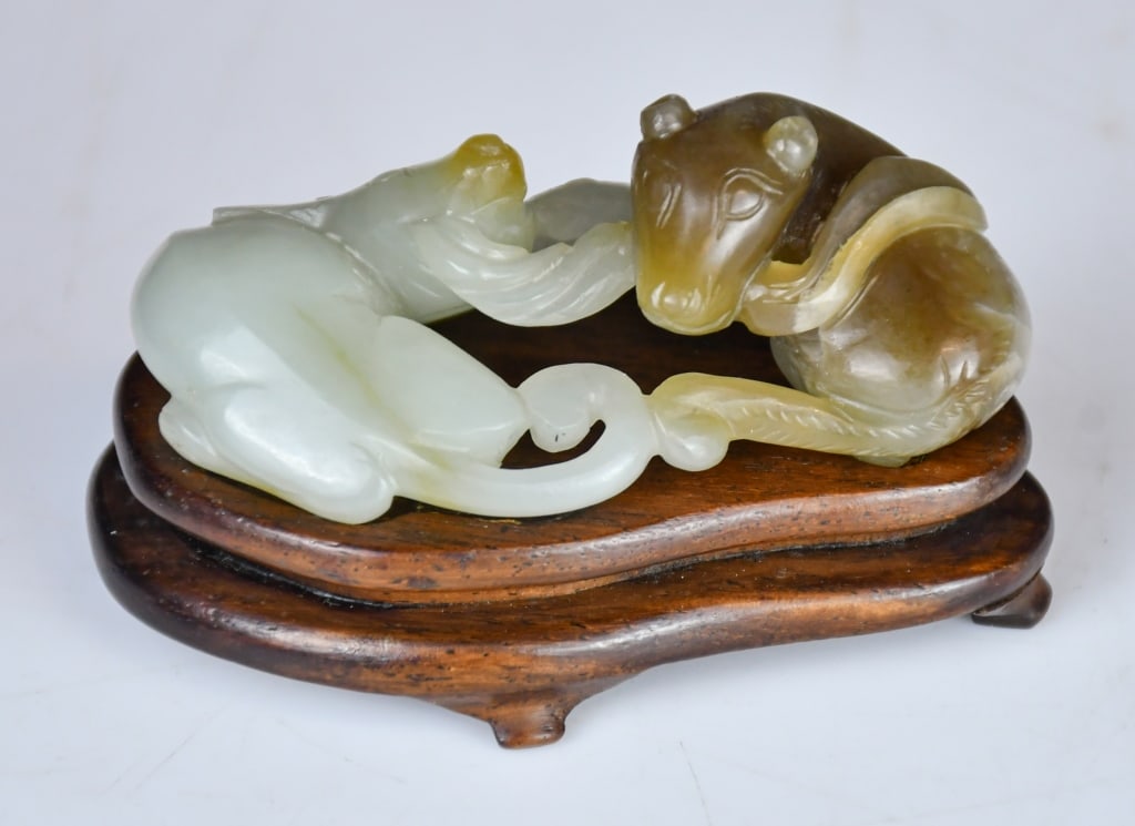A Carved Jade Double-Badgers Ornament Republican: A pale celadon jade carving depicting a pair of recumbent beasts with russet inclusions; comes with a wood stand. 8x4.3cm 民國 玉雕雙歡擺件