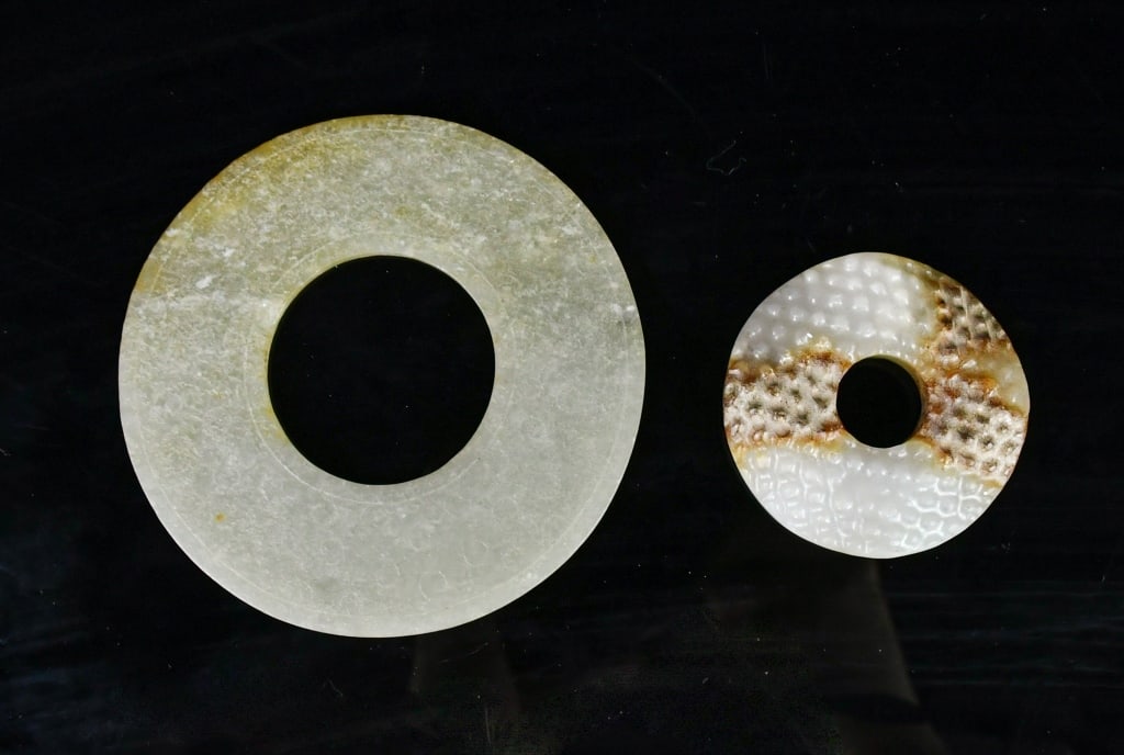 A Group of 2 Archaistic Jade Bi Discs: Comprising two jade bi discs, one worked from yellowish celadon jade, the other from white jade with russet inclusions, both with patterns on each side. Larger D: 9.5cm 