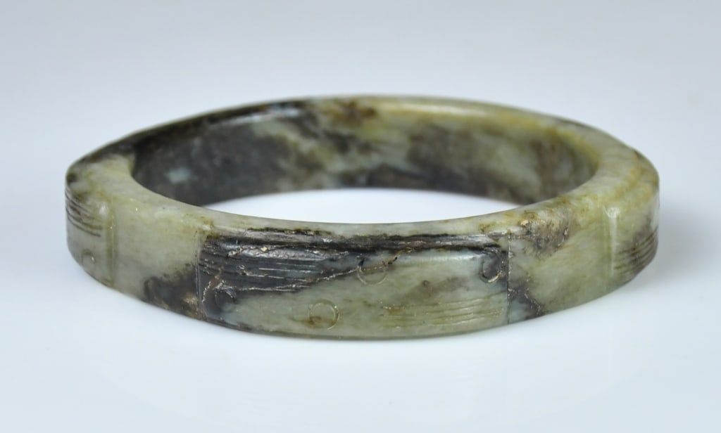 A Ming Style Jade Carved Bangle: A bangle of subtly faceted circular form, the edges gently rounded, the stone of celadon tone with dark brown, the exterior decorated with raised block-like reliefs carved with motifs. D: 8.4cm 