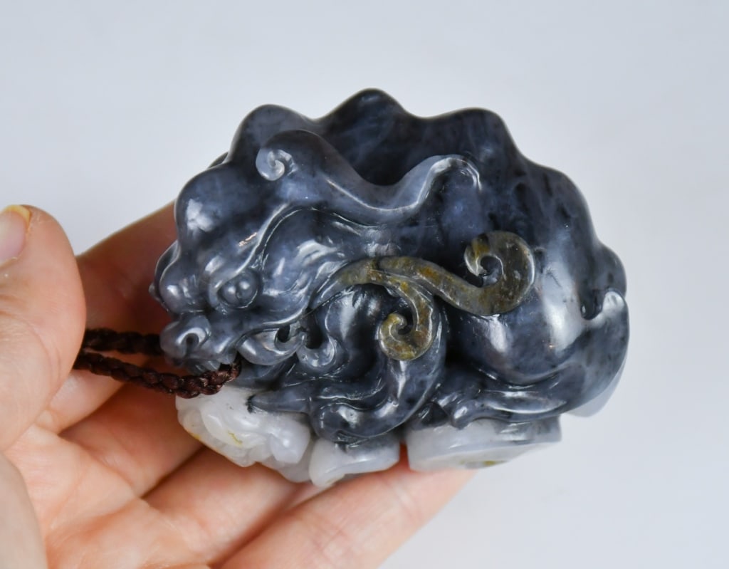 A Black & White Jade Caparisoned Beast 20thC: Sculpted in a recumbent posture with its head turned to the left, its jaws agape displaying sharp fangs. The body is rendered with stylized scrolls and cloud-like motifs along its flanks,feet step on