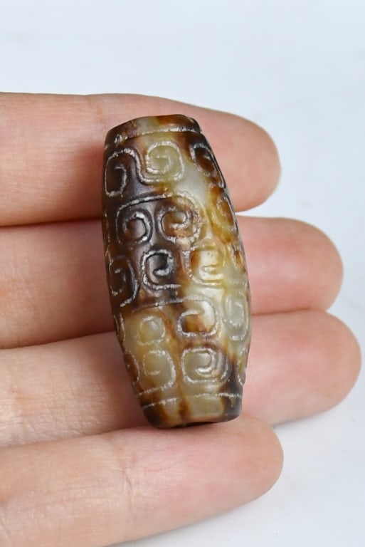 An Archaistic Carved Jade Tube: The hollow cylinder bead is of slightly swollen form, carved around the exterior in low relief with a continuous archaistic scroll design pattern. The stone is predominantly of a dark brown color,