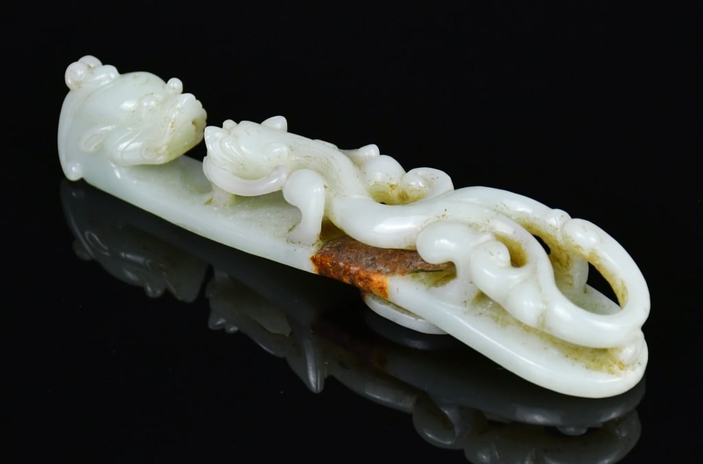 A Lrg Celadon Jade Dragon Belt Hook Qing: Finely carved with a chilong crawling towards the dragon-headed hook, the stone of pale celadon tone with russet inclusion. L: 14.5cm 清 青白玉大龍帶鉤