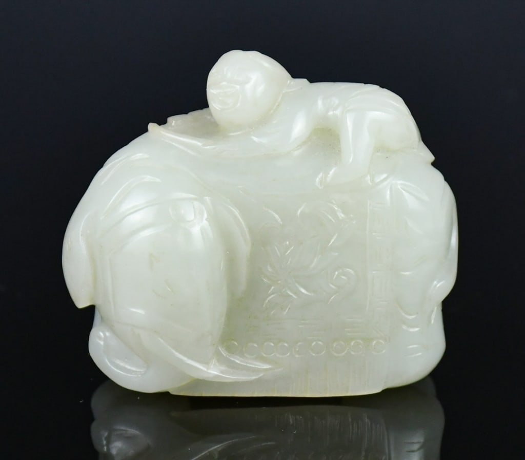 A Jade Carving of Elephant & Boy Ornament Republic: Depicts a young boy climbing atop the back of a recumbent elephant, the boy holding a cloth or branch that drapes over the animal's side. The elephant is carved with its head turned slightly to the ri