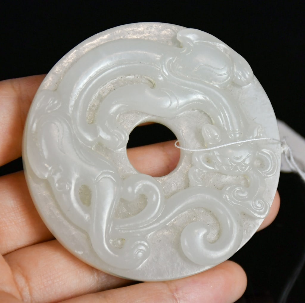 A White Jade Carved Chilong Bi Disc Qing: The circular white jade bi disc carved in relief with a chilong amidst scrolling cloud motifs, the reverse decorated with raised bosses, the stone of pale celadon-white tone. D: 5.9cm 清 白