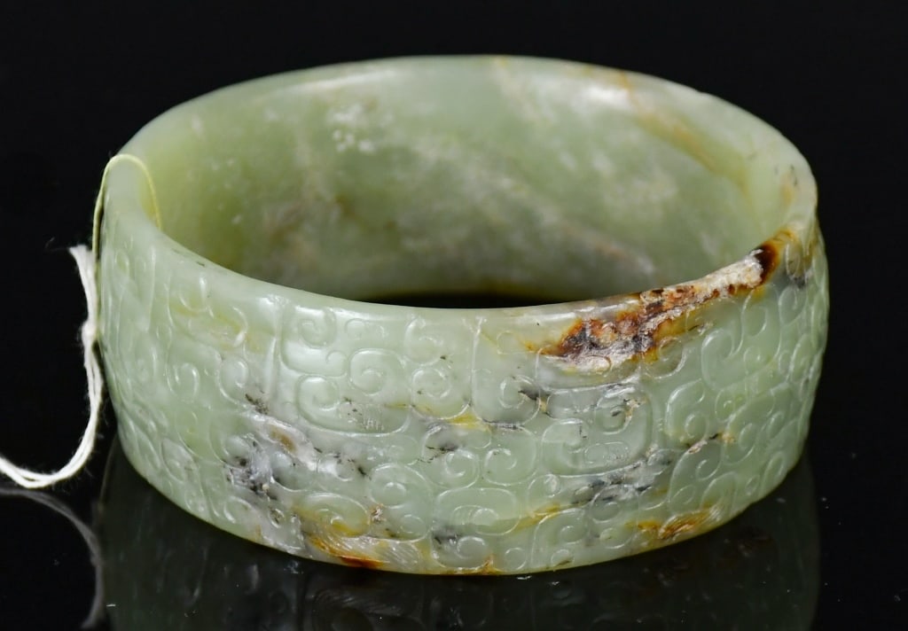 An Archaistic Celadon Jade Bangle: Worked from a celadon jade stone with russet inclusions, carved around the exterior with archaistic scroll patterns. D: 6.8cm 高古風格青玉雕雲紋&#
