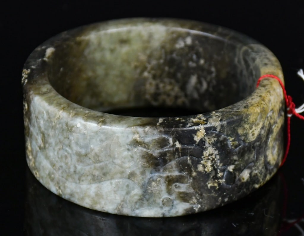 An Archaistic Jade Carved Bangle: Carved around the exterior with stylized dragon motifs, the stone of celadon tone with variegated dark and russet inclusions. D: 6.5cm 高古風格玉雕龍紋&